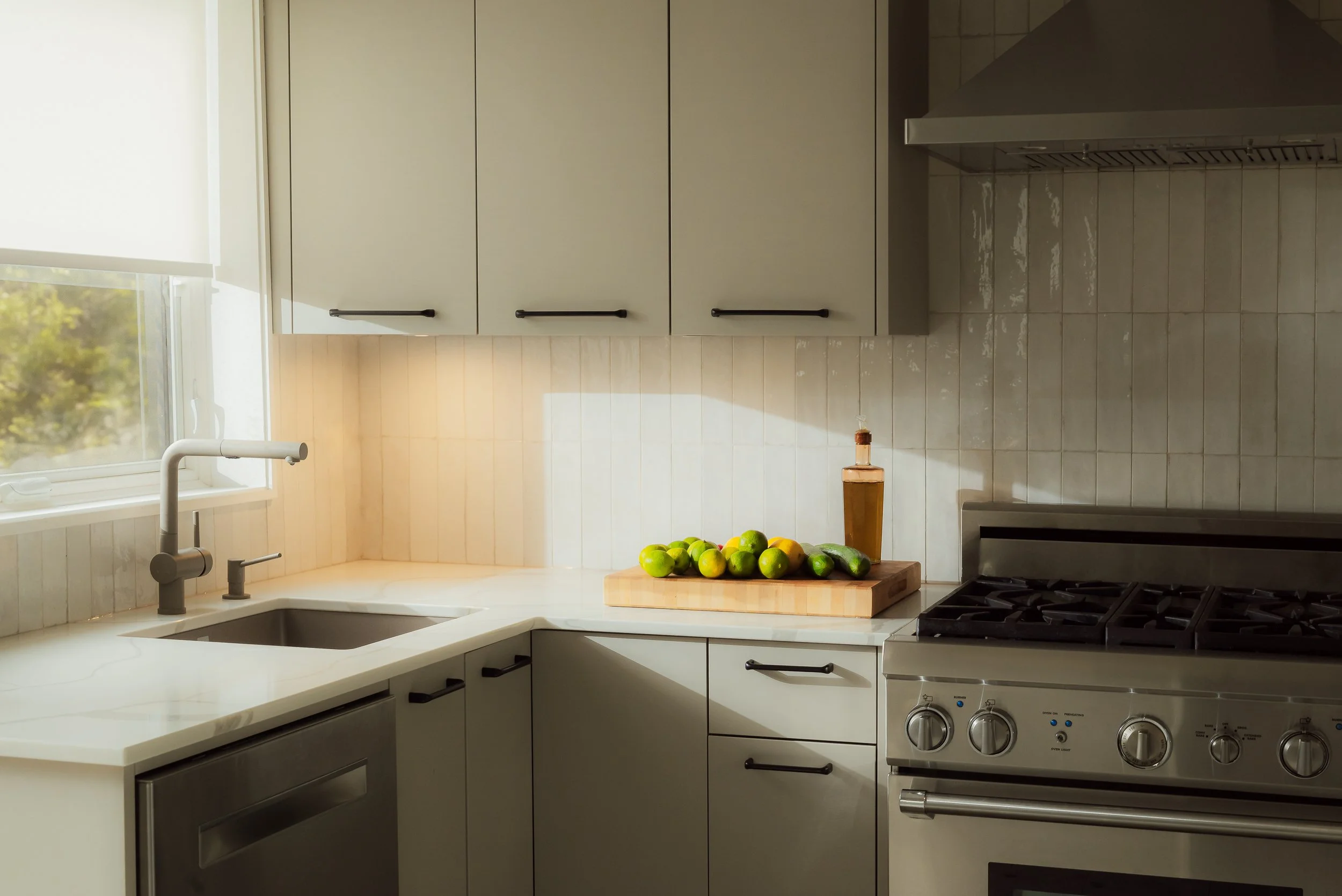 Kitchen with white cabinets, a window, a sink, a cutting board with limes, lemons, and a bottle of oil, and a stainless steel stove.