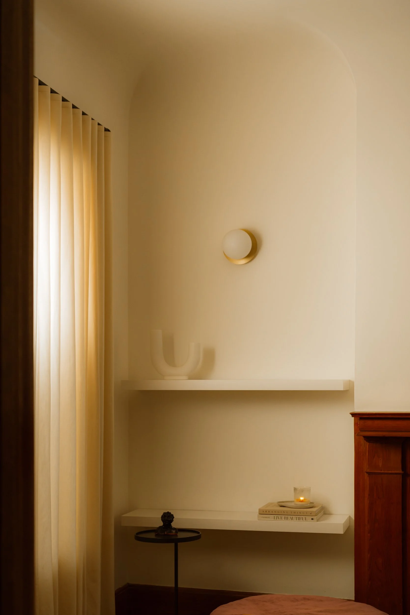 A corner of a living room with a window covered by beige curtains, a wall-mounted light fixture, a white floating shelf with decorative items, and a small round black side table with a decorative object. There is also a candle on a stack of books and a wooden piece of furniture on the right.