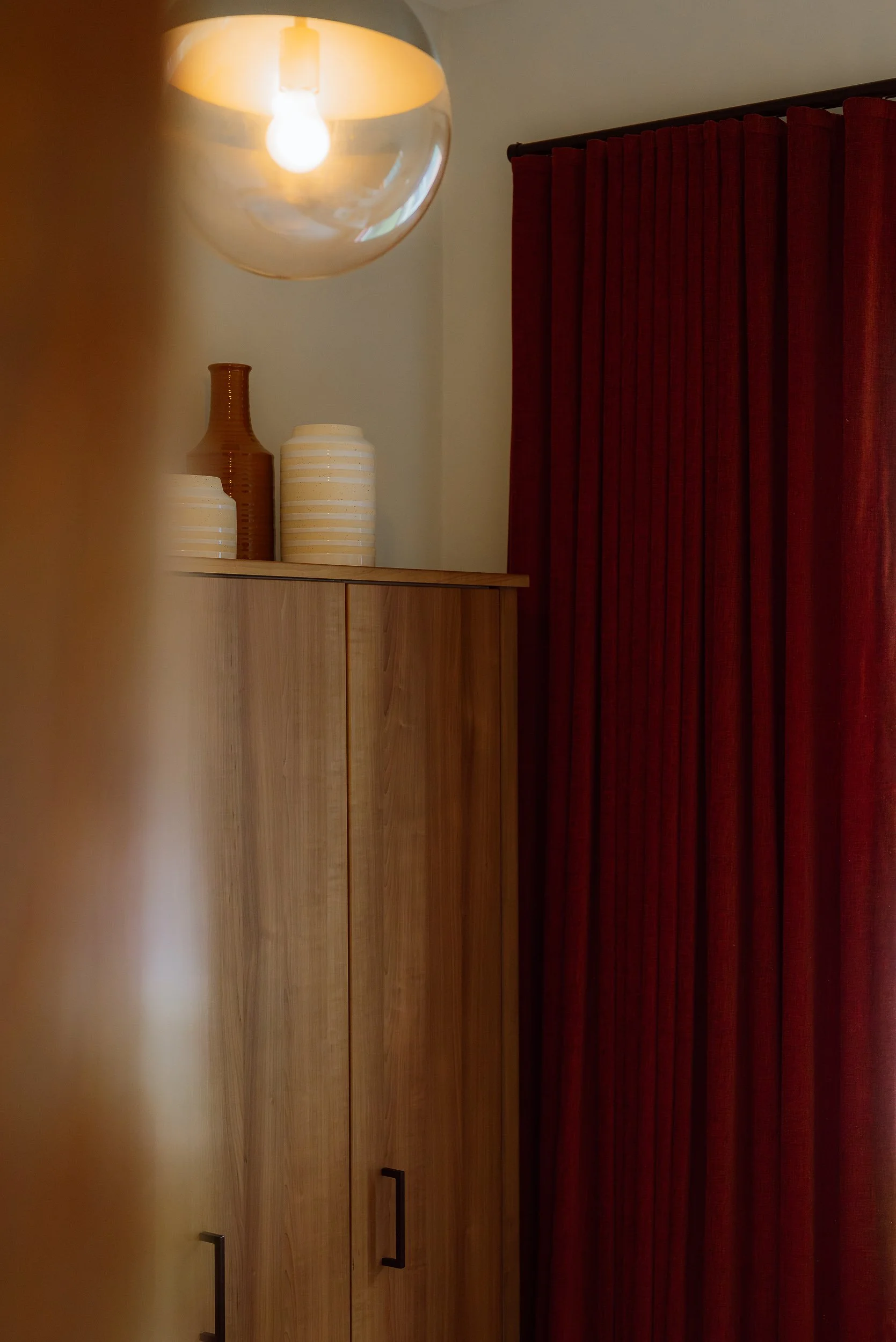 Part of a room with a wooden cabinet, red curtains, a hanging ceiling light, and decorative vases on top of the cabinet.