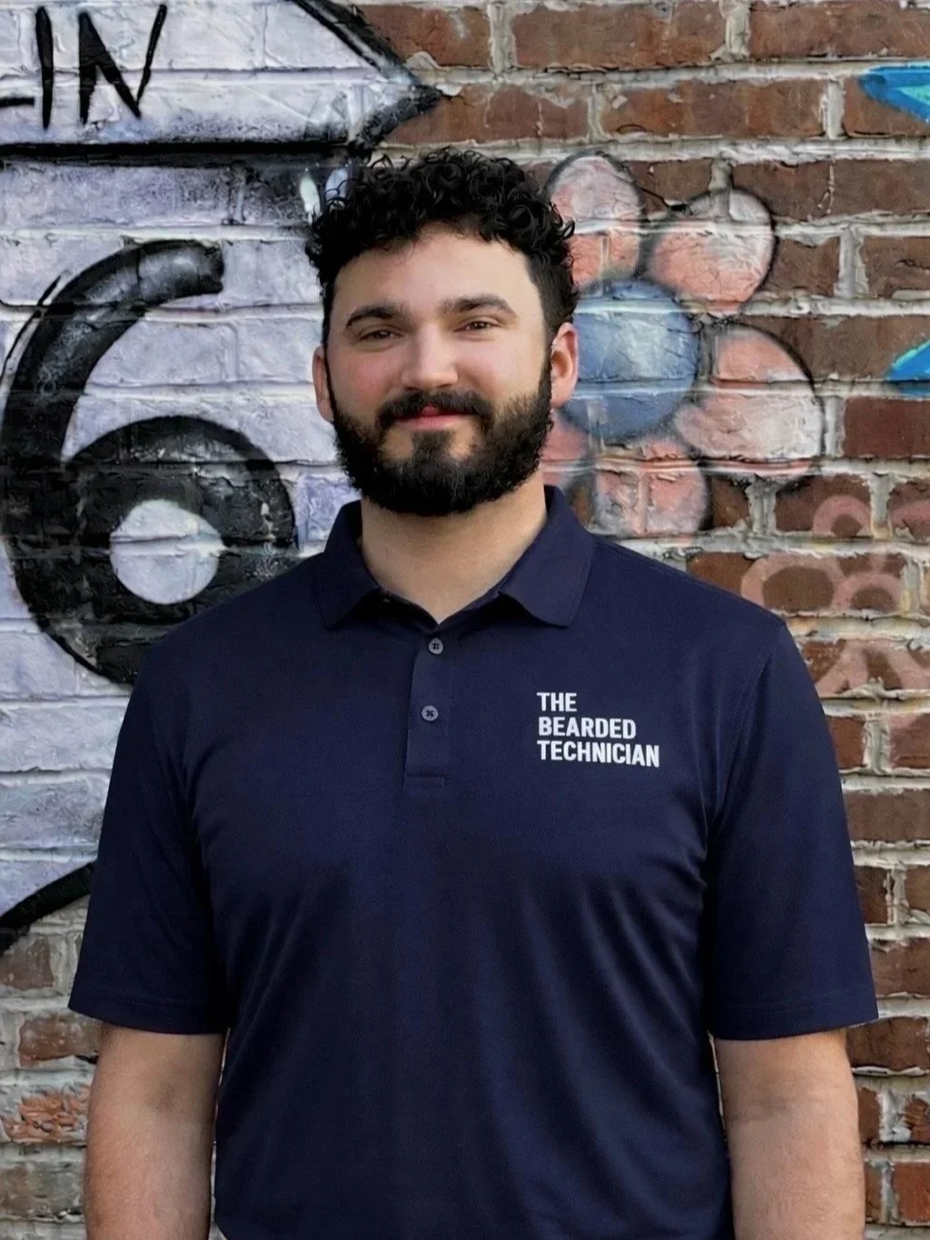 Man with dark curly hair and beard standing in front of a brick wall with graffiti, wearing a navy polo shirt that reads "The Bearded Technician."