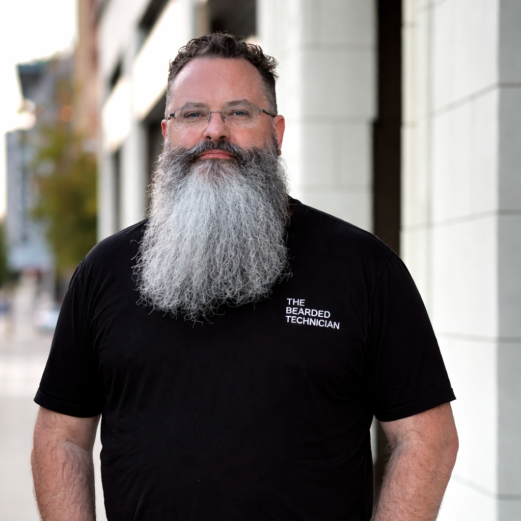 A bearded man wearing glasses and a black T-shirt that reads 'The Bearded Technician' standing outdoors with a blurred city background.