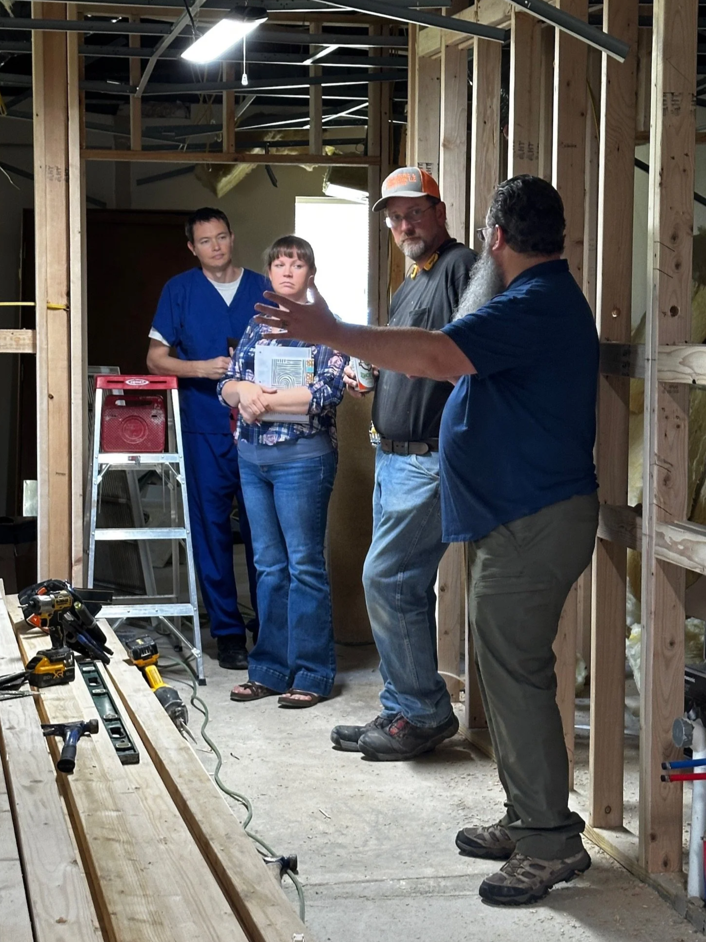 A group of four people inside a house under construction. One person, standing on the right, is talking and gesturing with his hands. Two other people, a man and a woman, are listening and observing. The fourth person, a man in blue scrubs, is standing in the background. The room has exposed wooden framing, a ladder, and power tools on a wooden workbench.