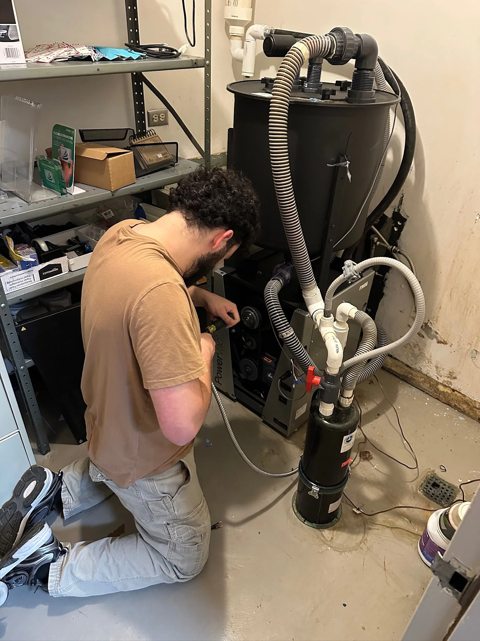 A man kneeling on the floor, working on an HVAC heating and cooling system inside a utility room with shelves and equipment.