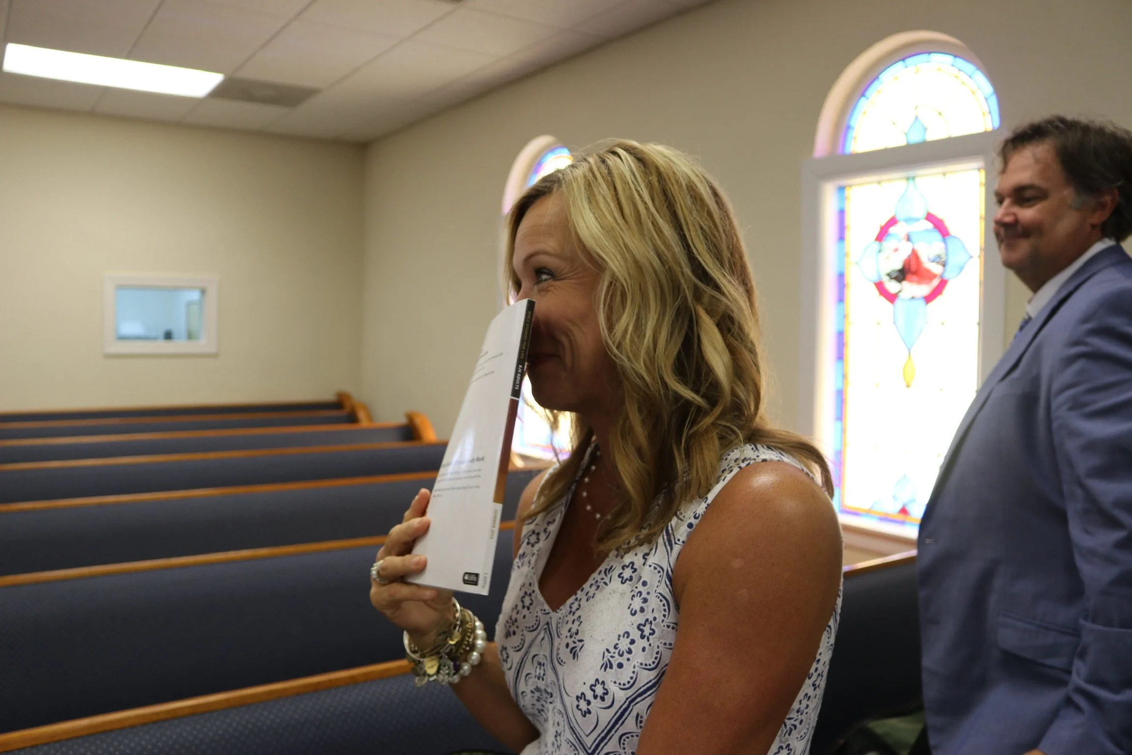 A woman smiling and holding a book up to her face in a church with stained glass windows, a man standing behind her, and empty pews.