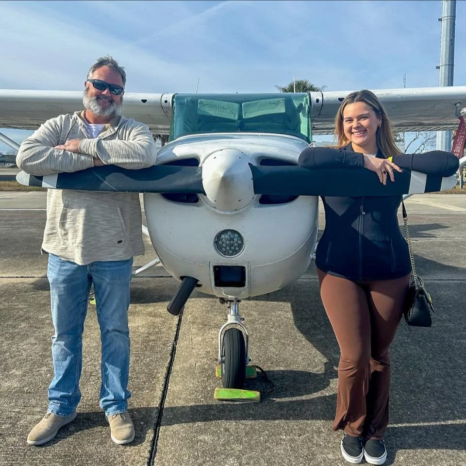 A man and a woman are standing on either side of a small airplane, smiling, with arms resting on the plane's wings. The scene is outdoors on a sunny day.