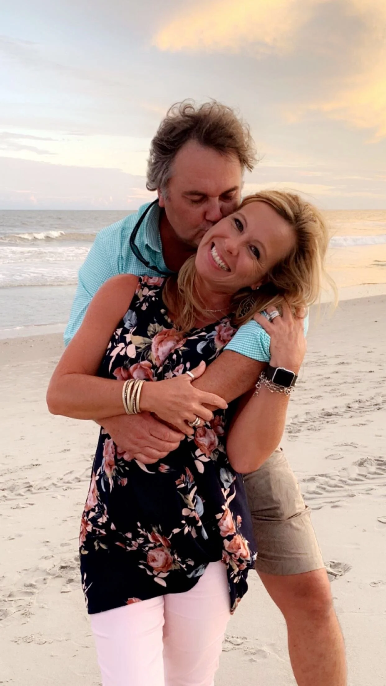 A happy couple hugging on the beach during sunset, with the man kissing the woman on the cheek, both smiling joyfully.