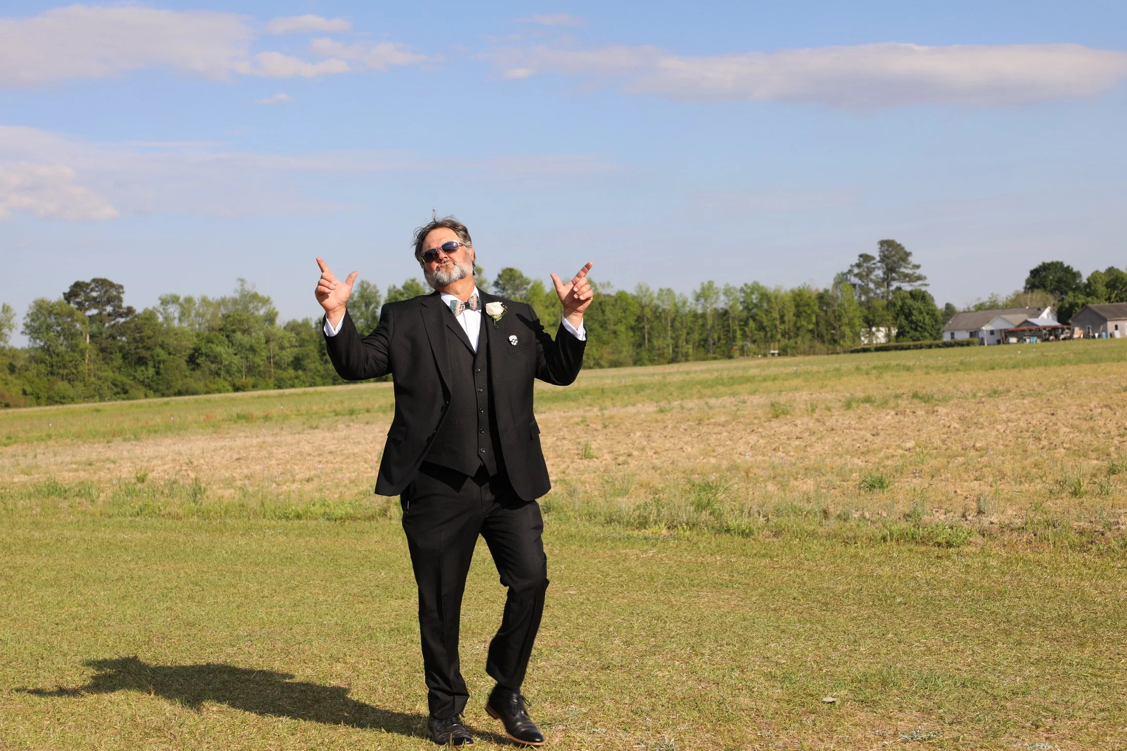 A man in a black tuxedo with sunglasses and a boutonniere on his lapel, standing outdoors on a grassy field with a blue sky and clouds, striking a playful pose with both thumbs up.