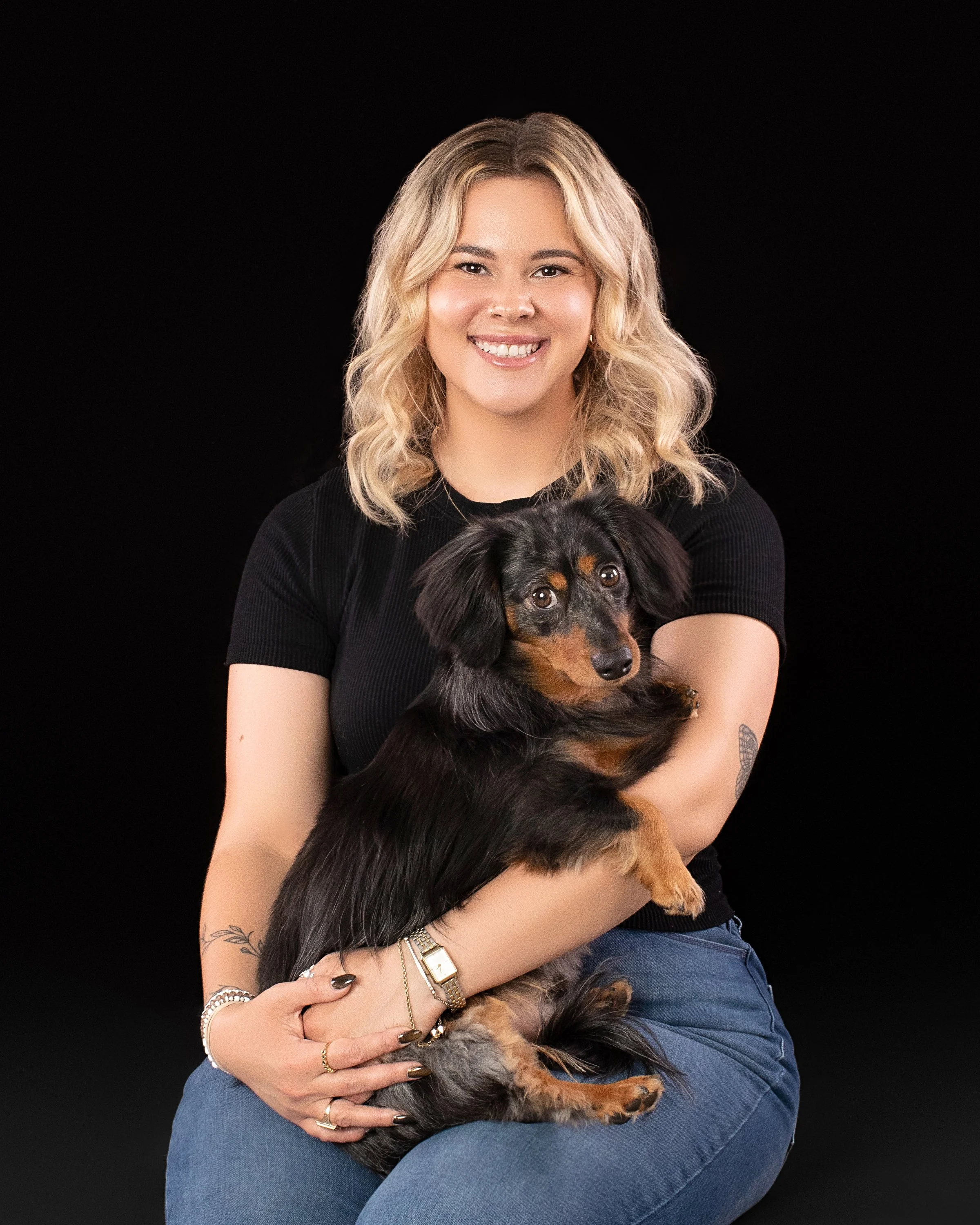 A woman with blonde, wavy hair smiling and holding a small black and brown long-haired dachshund against a black background.