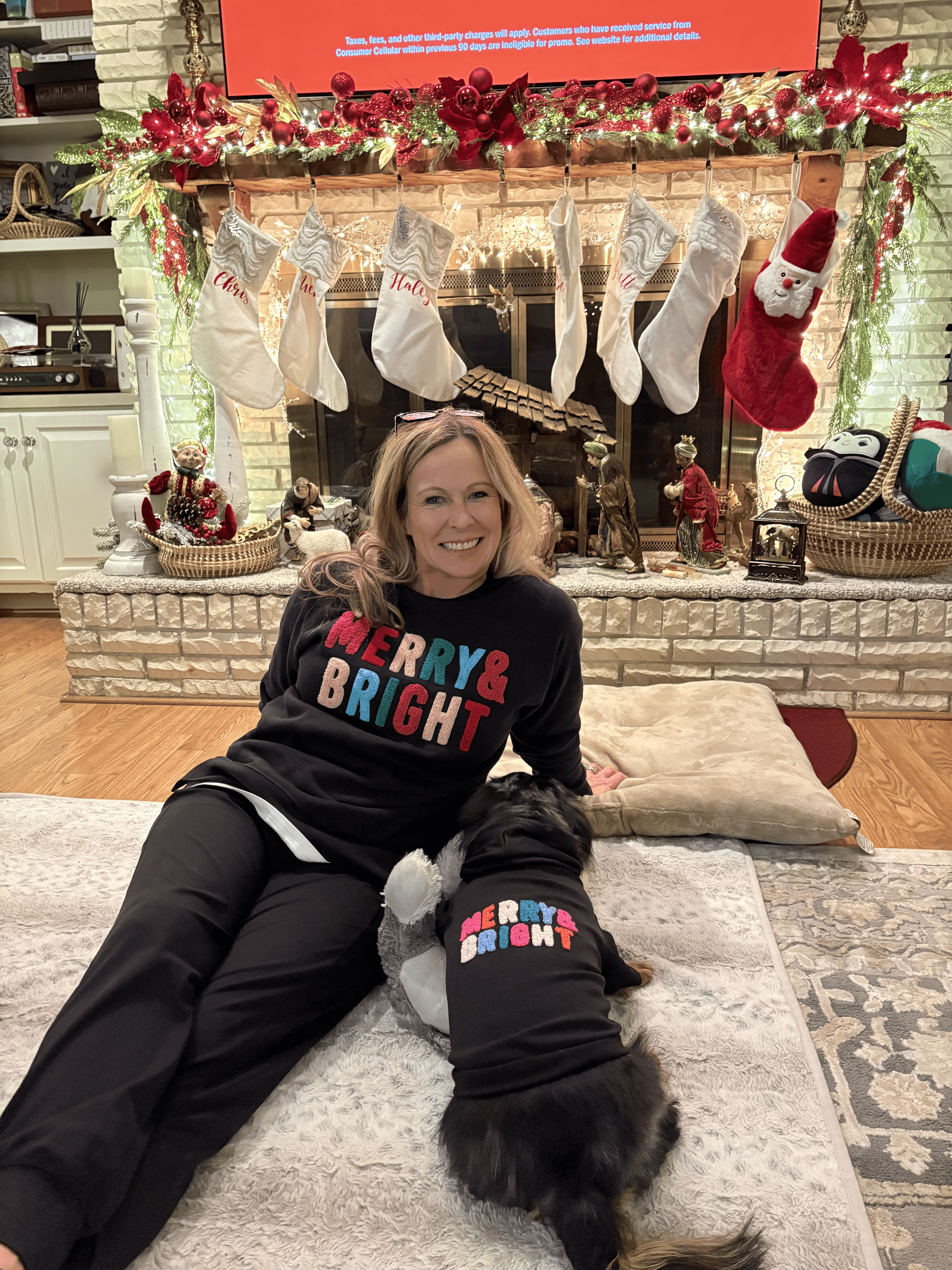 A woman and a dog wearing matching "Merry & Bright" sweaters sit on the floor in front of a decorated fireplace with Christmas stockings and ornaments. The woman is smiling, and the dog is lying next to her.