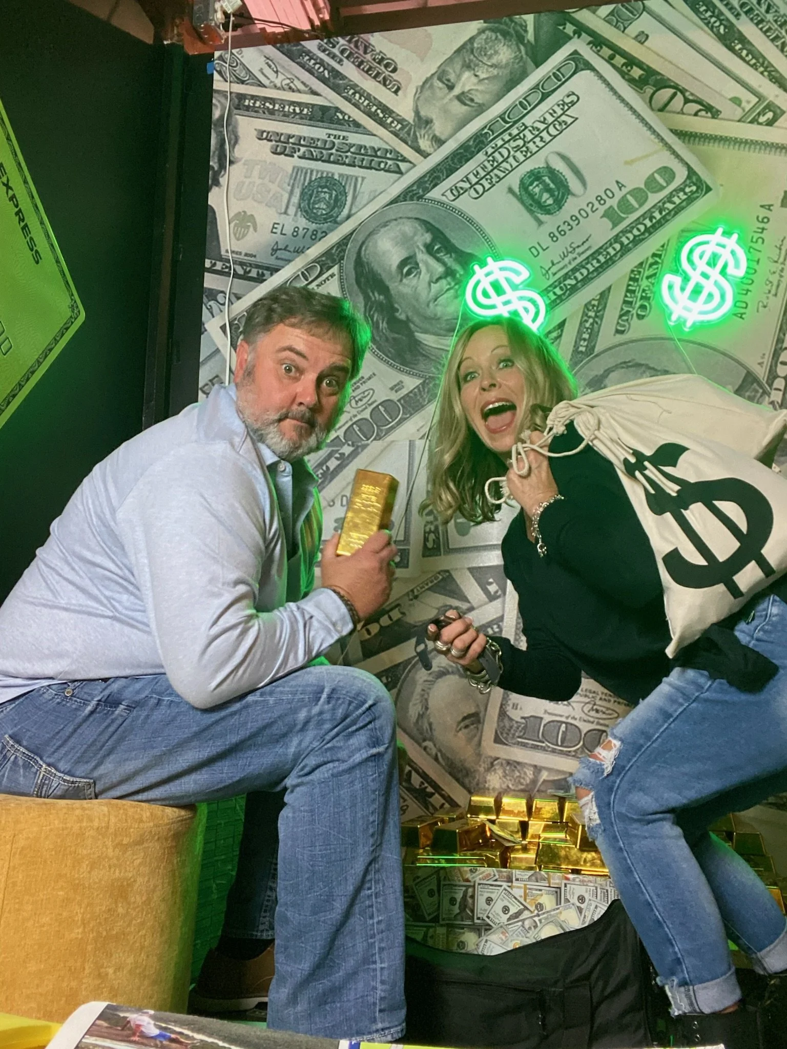 Two people inside a money-themed room with $100 bills on the wall. The man on the left holds a gold bar, and the woman on the right holds a bag with a dollar sign. There are neon dollar signs above their heads and piles of gold bars and cash on the floor.