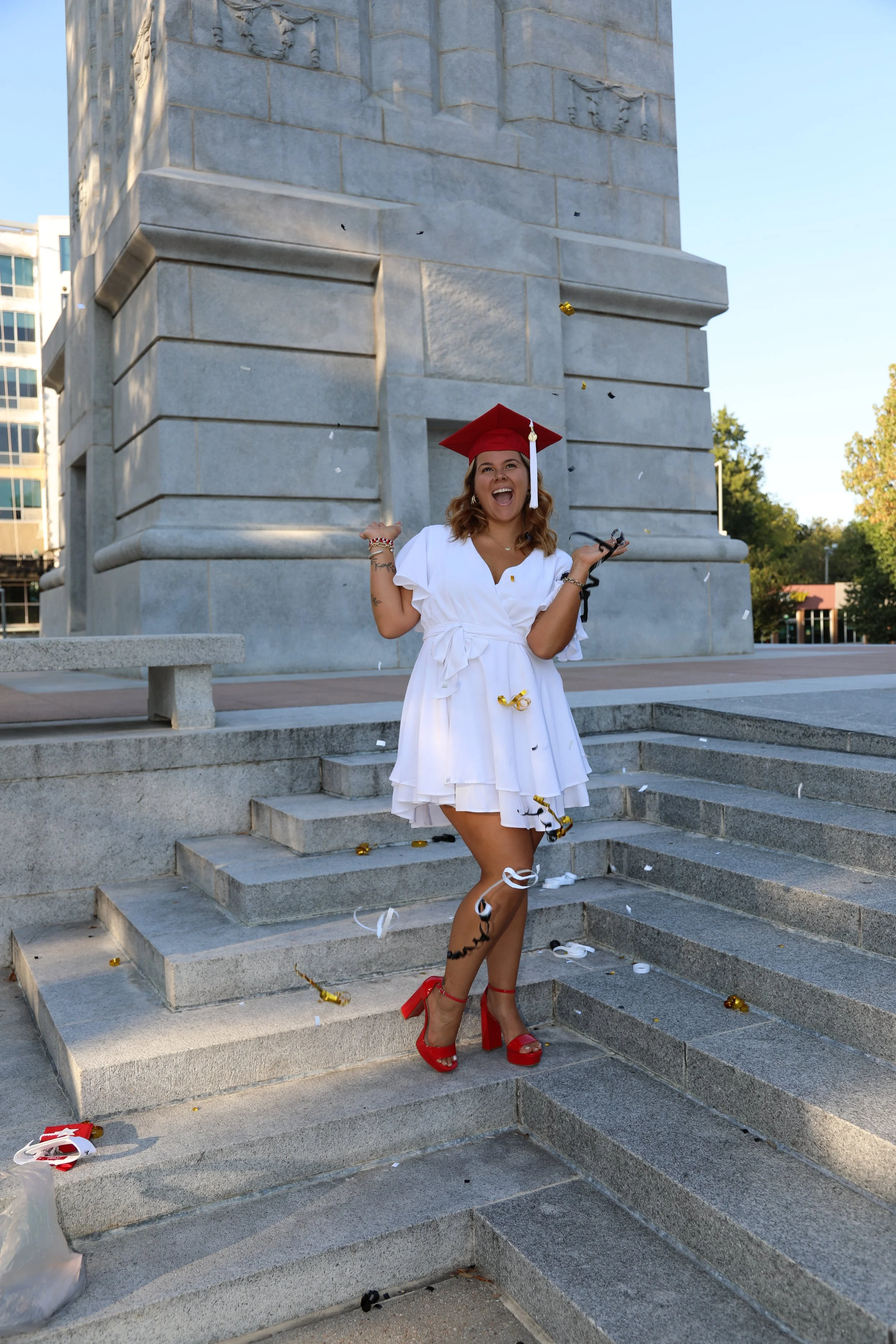 Young woman in a white dress and red high heels celebrating with a red graduation cap, standing on steps in front of a stone monument with scattered confetti and streamers.