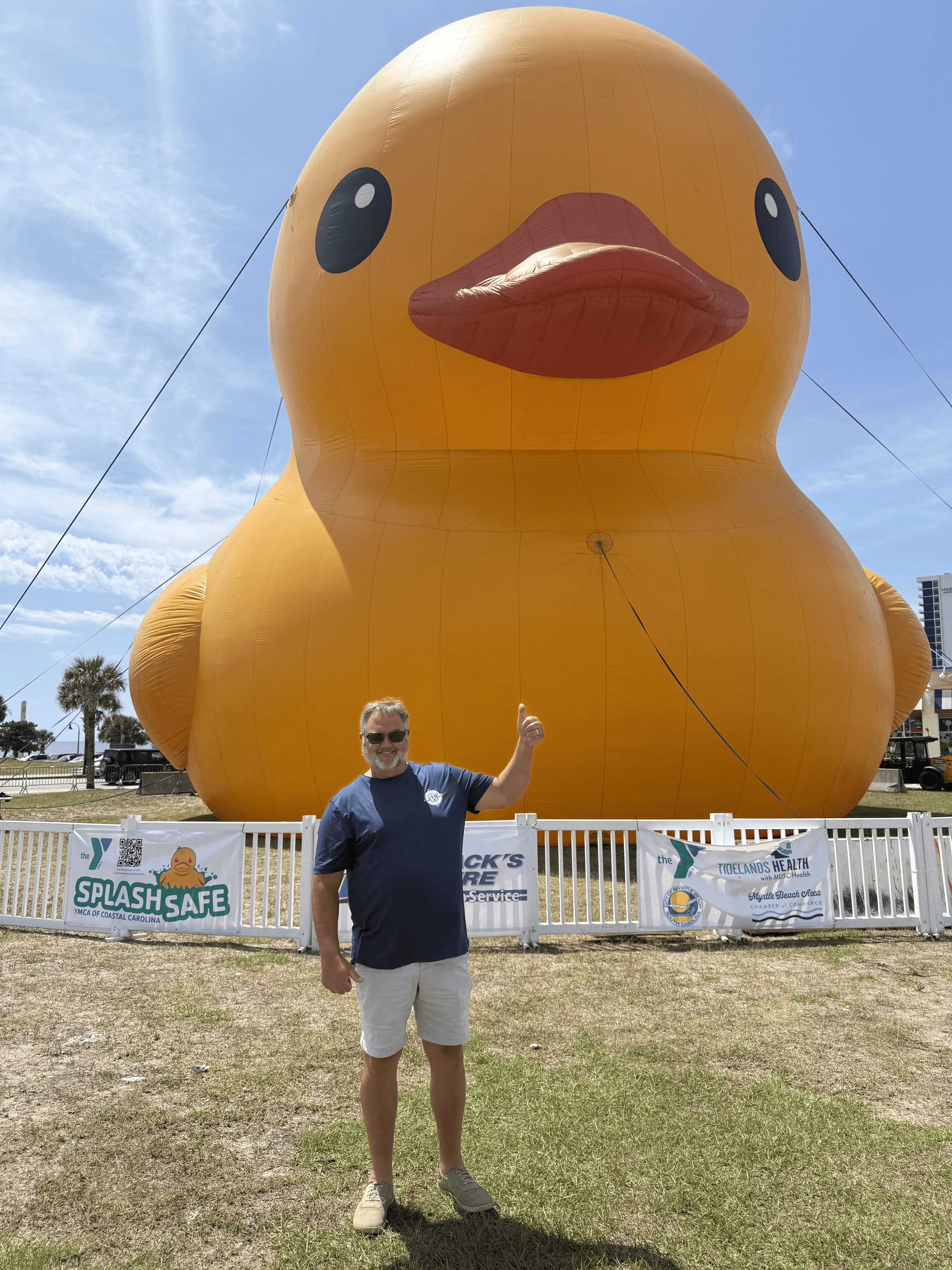 A man wearing sunglasses, a blue T-shirt, and khaki shorts standing outdoors, giving a thumbs-up in front of a giant inflatable yellow rubber duck. The duck is near a white fence with banners, and the sky is blue with some clouds.