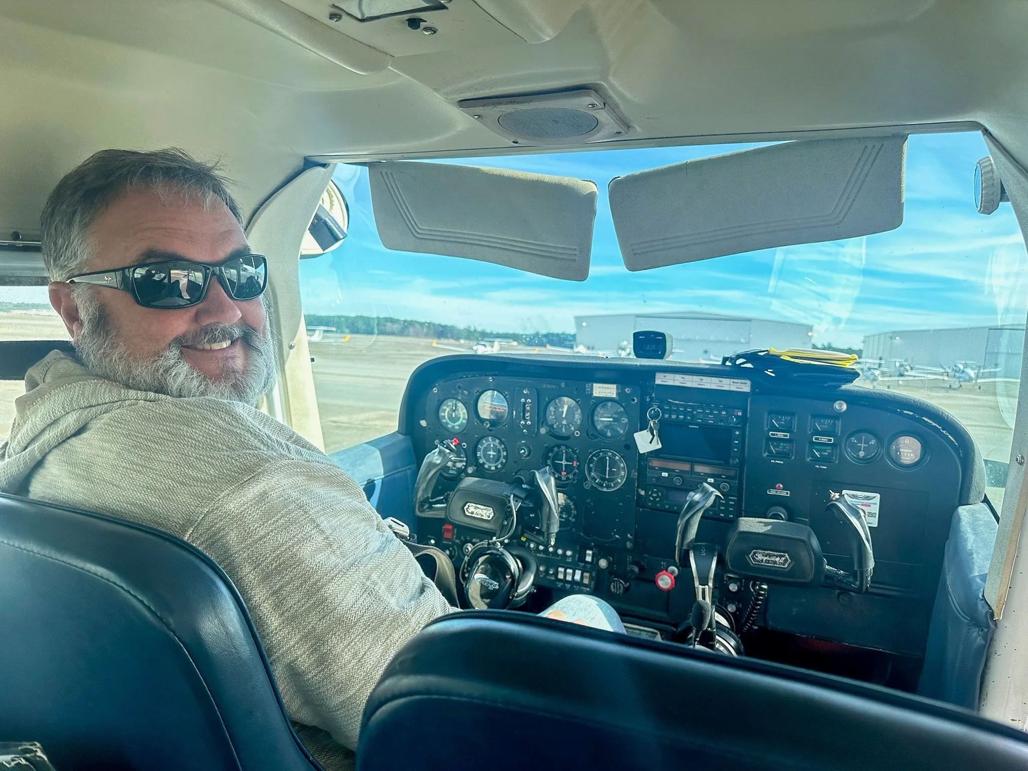 A man with grey hair and beard wearing sunglasses, sitting in the cockpit of a small airplane at an airport, smiling at the camera, with aircraft and runway visible through the front window.