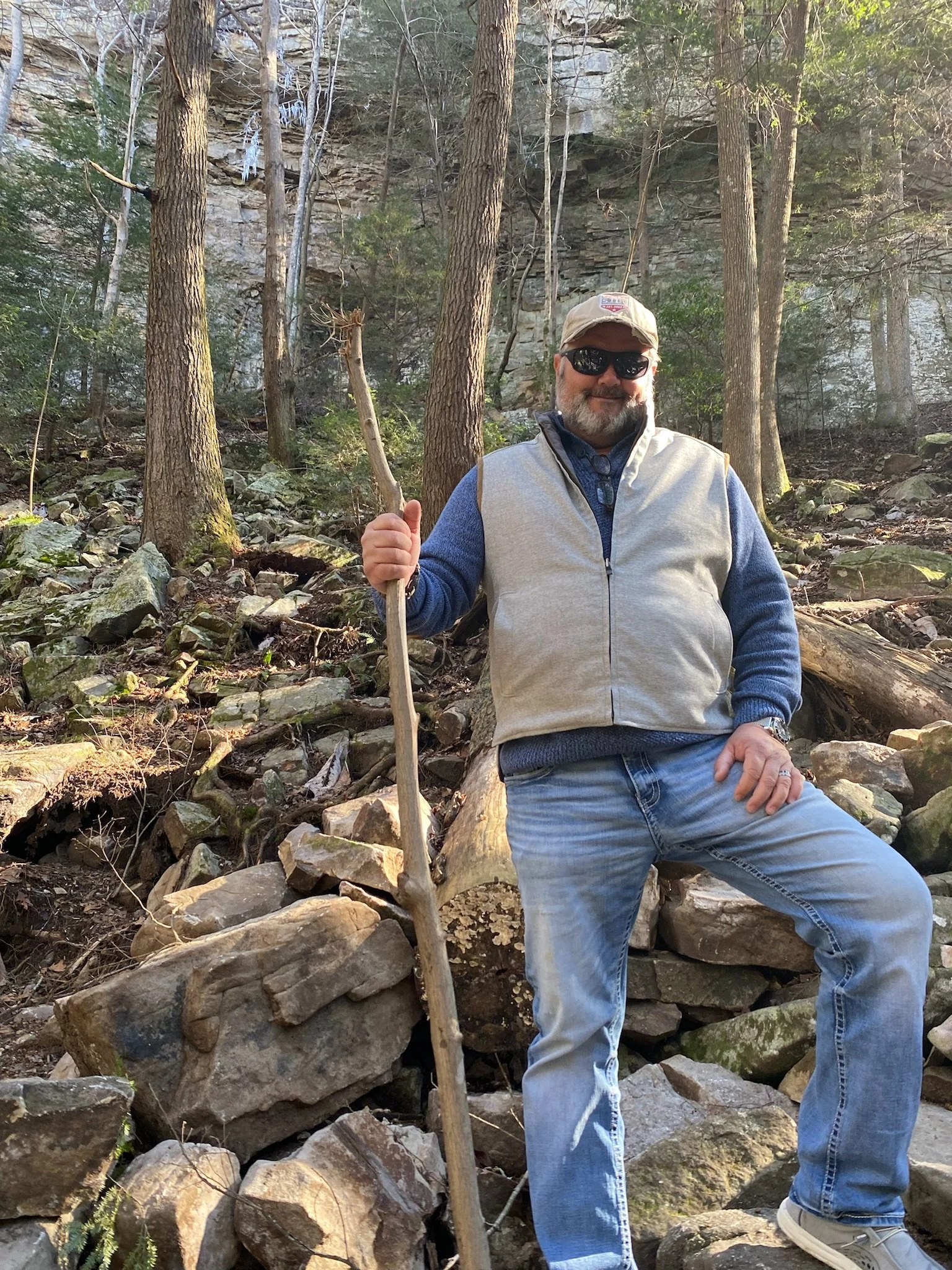 A man with sunglasses, a beige cap, a blue sweater, and a gray vest standing outdoors in a rocky wooded area, holding a long walking stick.