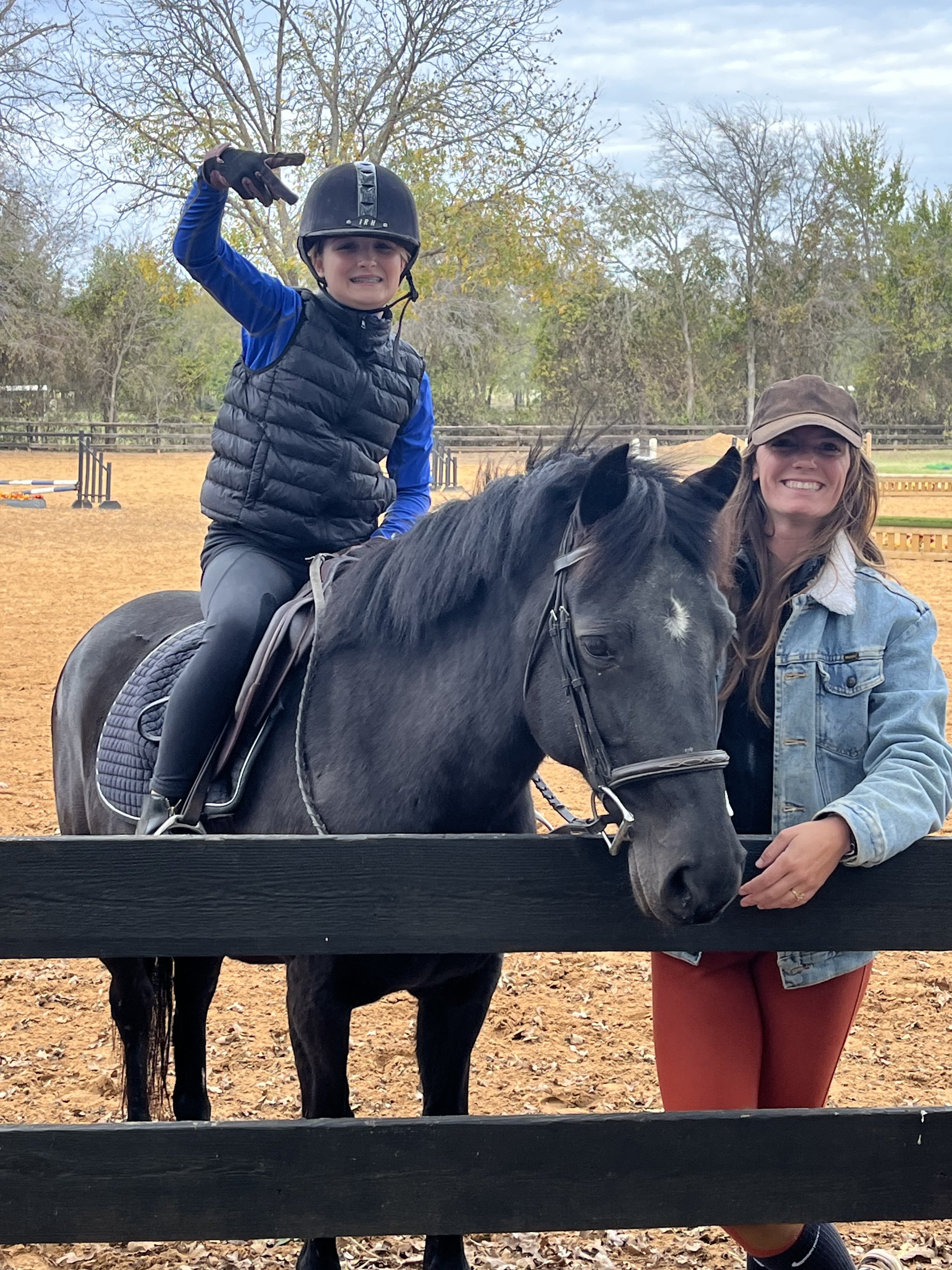 Young girl wearing a helmet and vest riding a black pony, raising a hand and making a peace sign. Trainer standing beside the horse, smiling, touching its neck. The scene takes place in an outdoor riding arena with trees and jumps in the background.