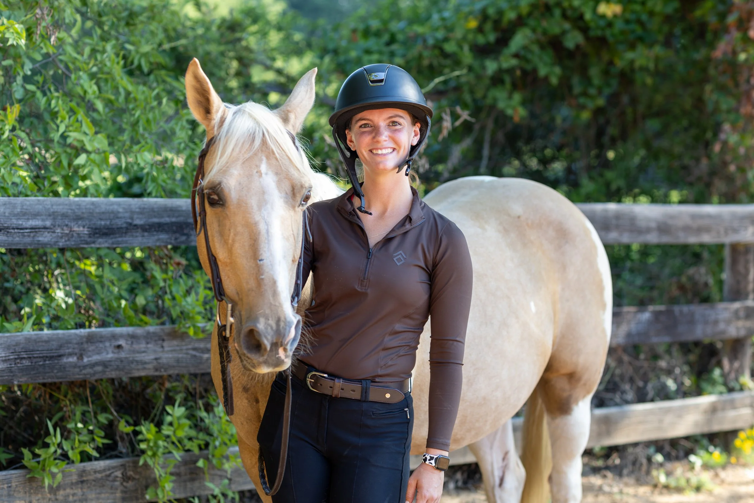 A smiling woman wearing a helmet and riding outfit standing next to a light-colored horse with a wooden fence and greenery in the background.