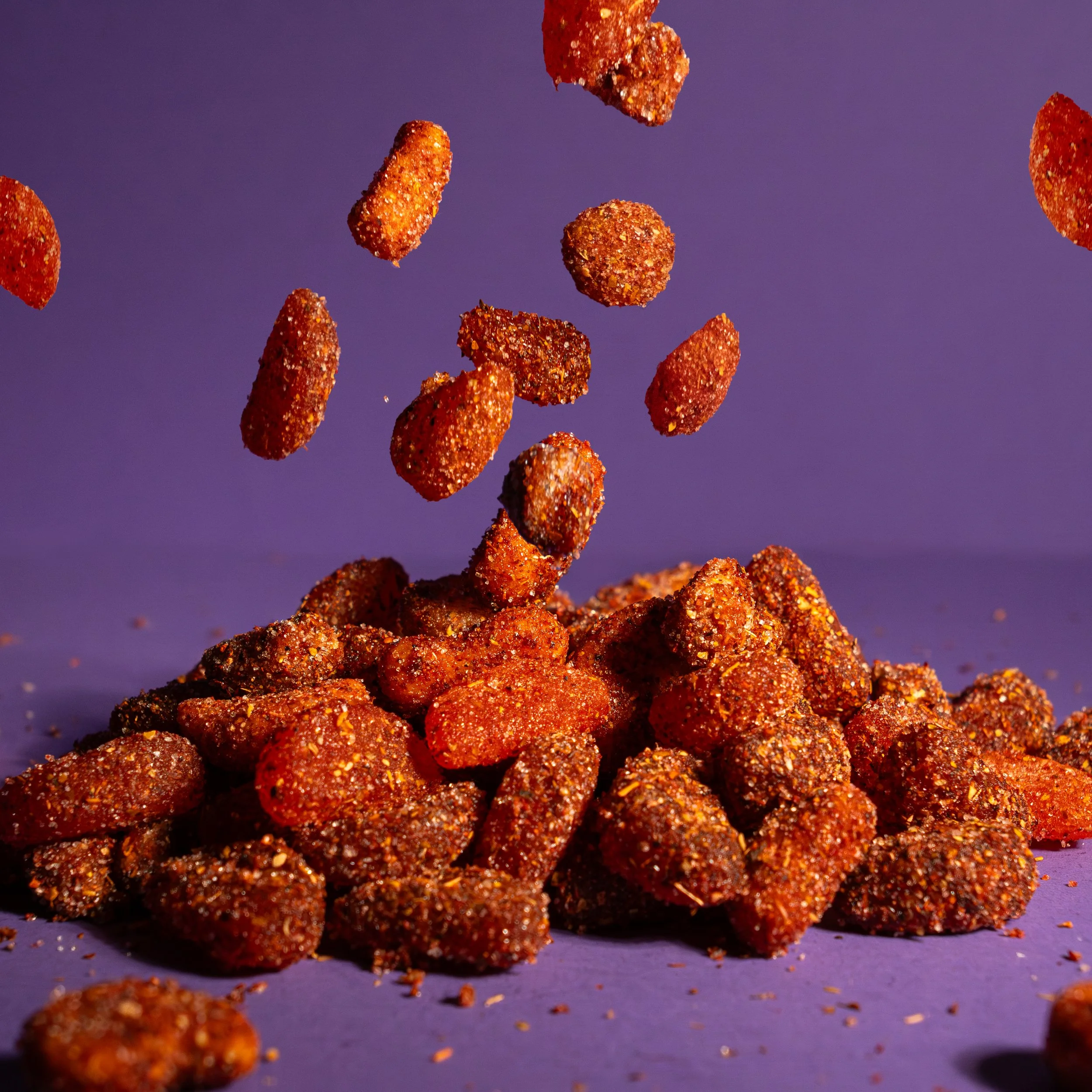 Trozos de gomitas de sabores mango piña y naranja cubiertos de mezcla de chiles en polvo a granel, cayendo sobre un fondo morado.