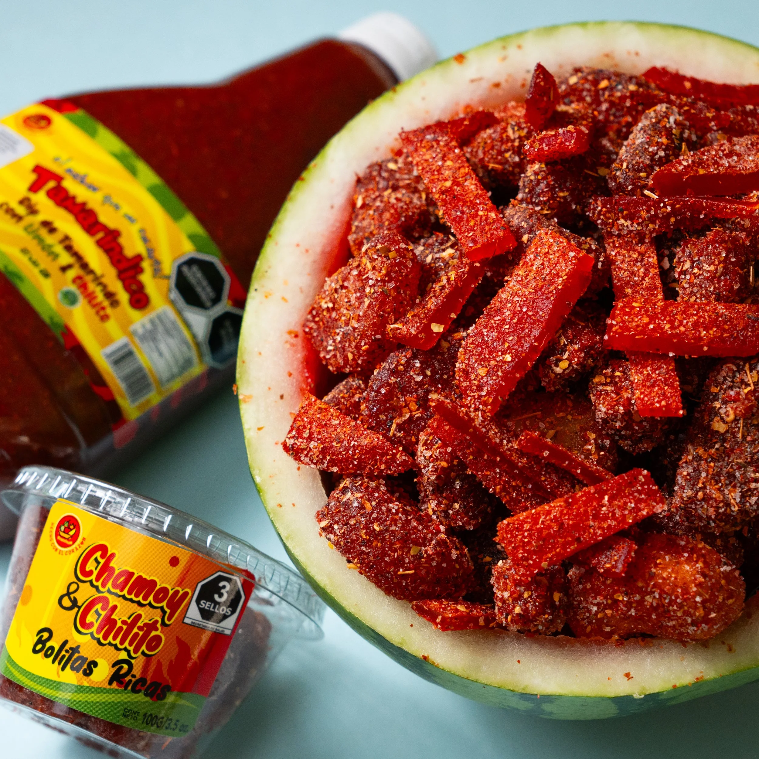 Sandía rellena de chiles en polvo, junto a botellas de dip salsa chamoy picante y un paquete de bolitas de fruta enchiladas