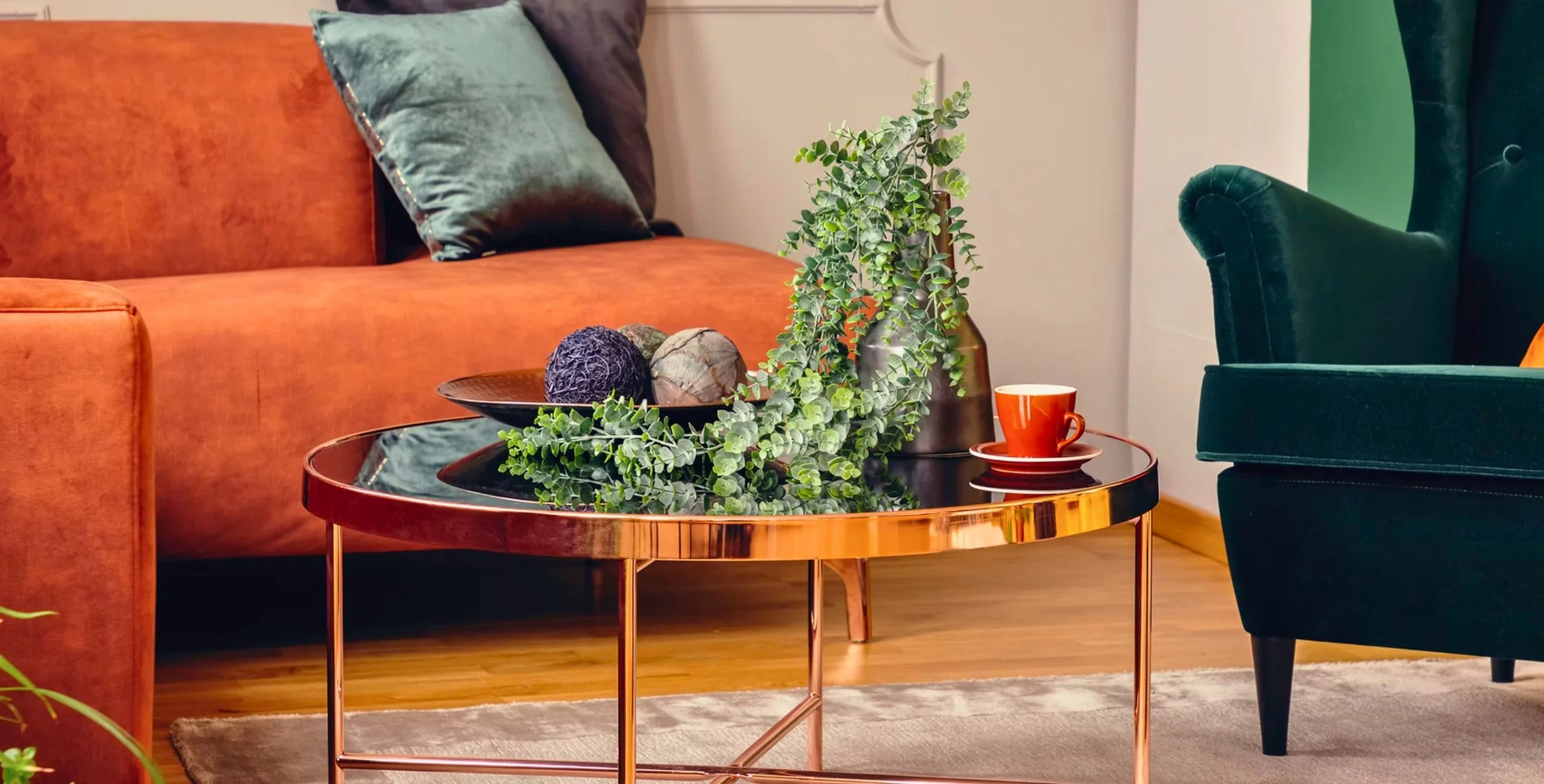 Living room with an orange velvet sofa, a green velvet armchair, a round black and copper coffee table with decorative items including a plant, stones, and a mug, and a wooden floor.