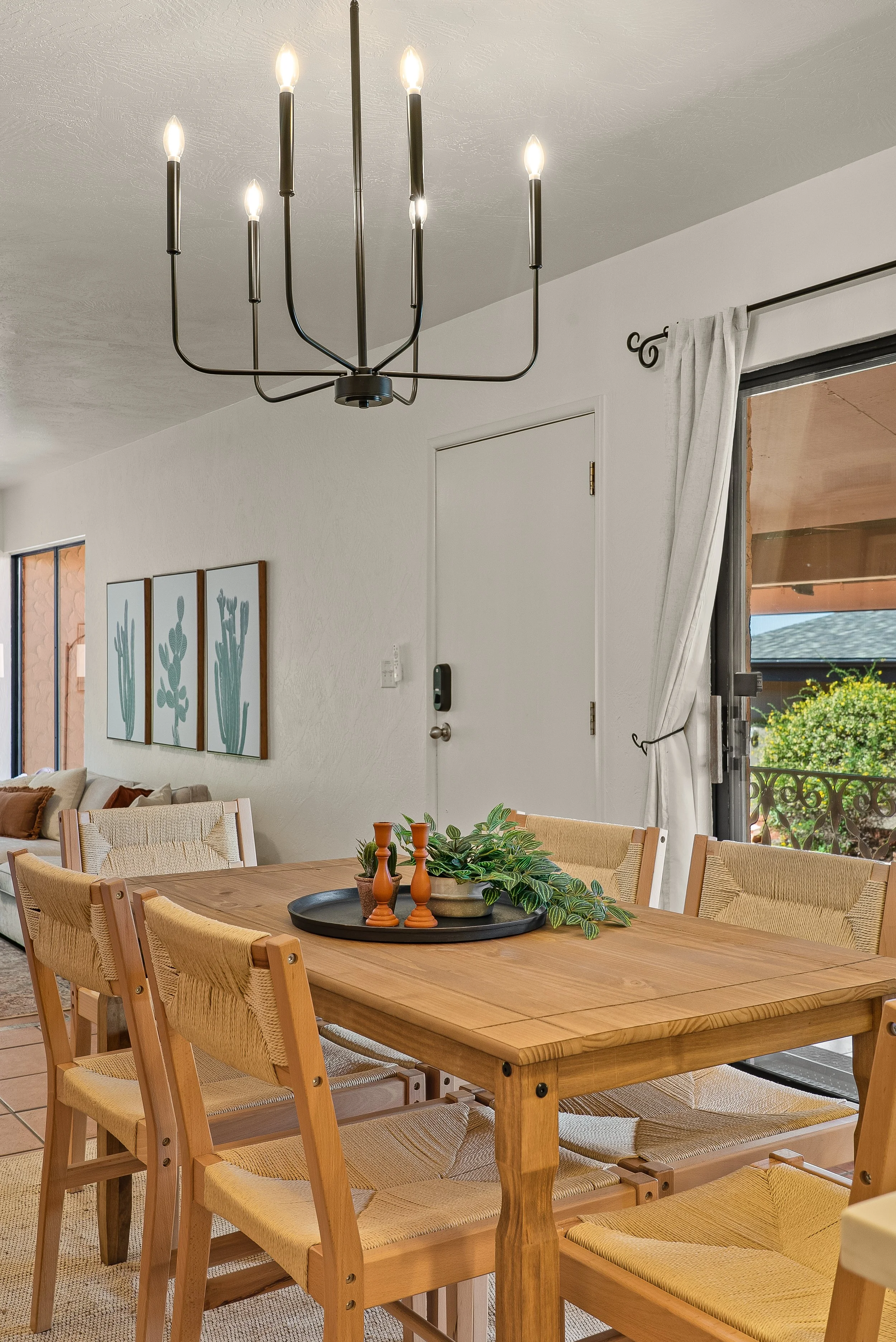 A dining room with a wooden table, beige chairs, a black chandelier with candle-shaped lights, sliding glass door with white curtains, and wall art of three cacti.