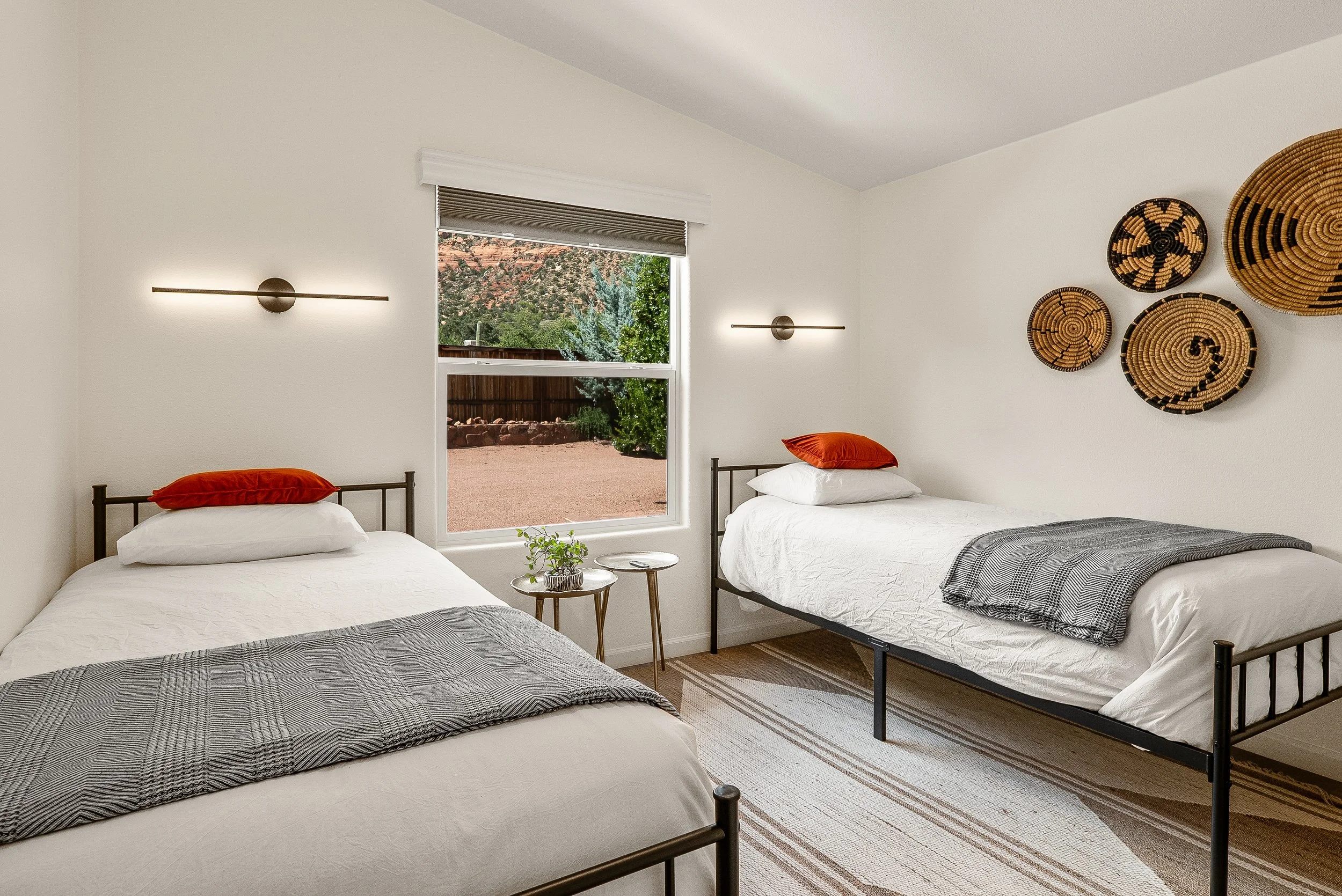 Bedroom with two beds, each with white bedding, orange pillows, and gray blankets, wooden side table with green plant, window showing desert landscape with trees and rocks, wall decor with five woven baskets, and modern wall lights.