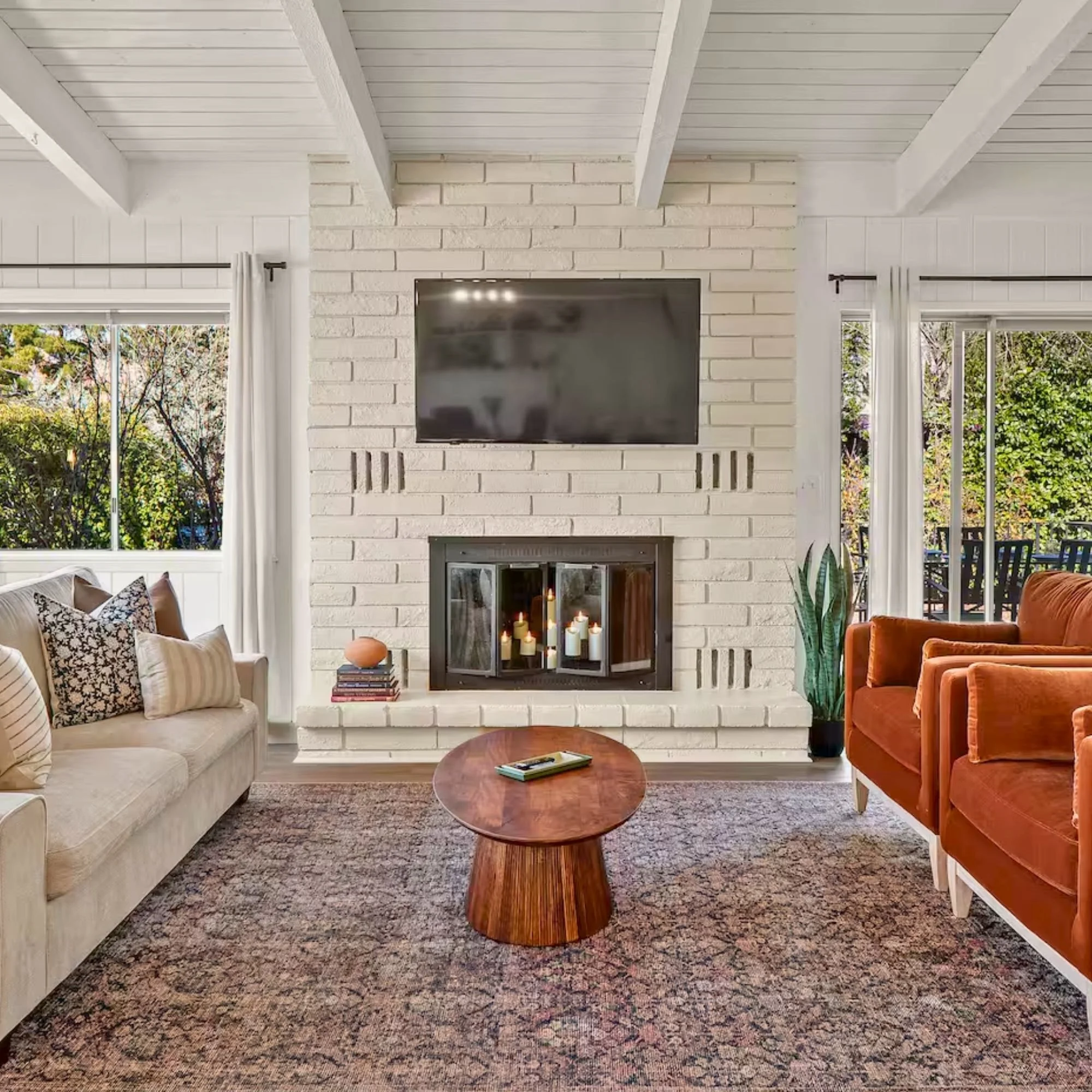 Living room with white brick fireplace, mounted flat-screen TV, glass firewood door with candles inside, beige and orange sofas, wooden coffee table, pattern rug, large windows with curtains, and greenery outside.