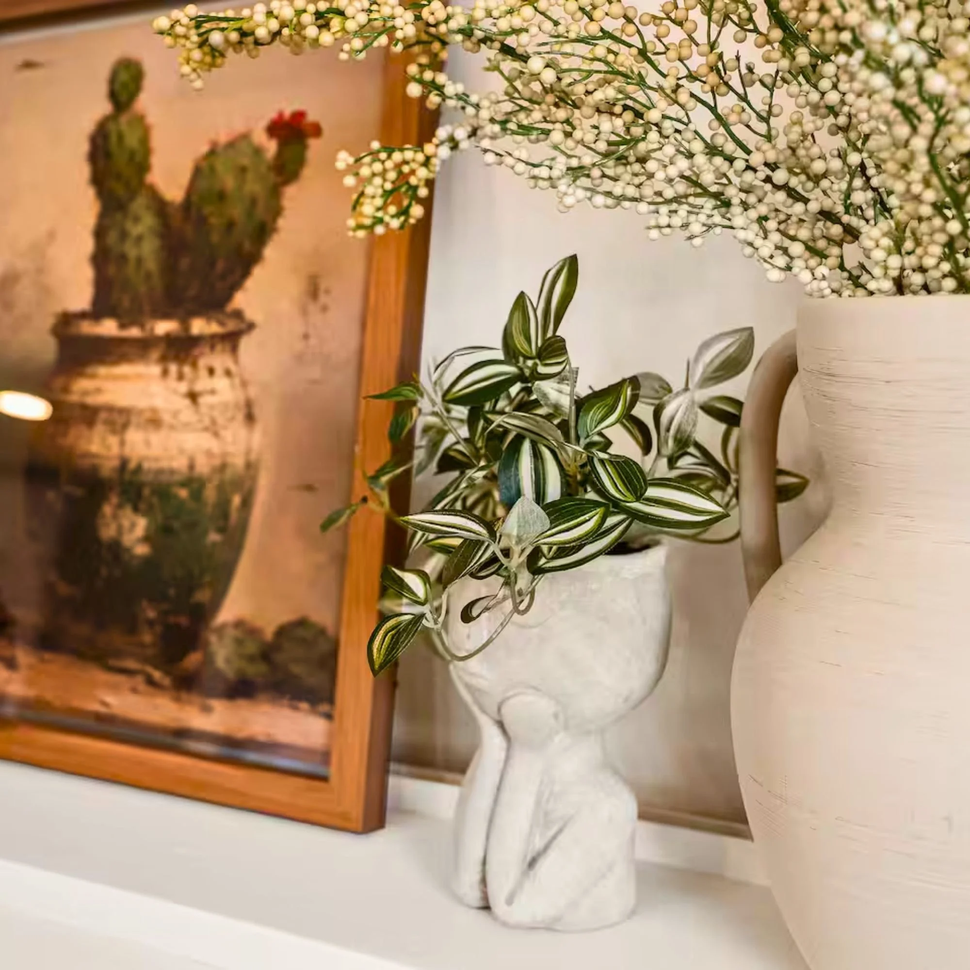 Decorative indoor arrangement with potted plants, a photograph of a cactus in a pot, and white vases with white flowers on a shelf.