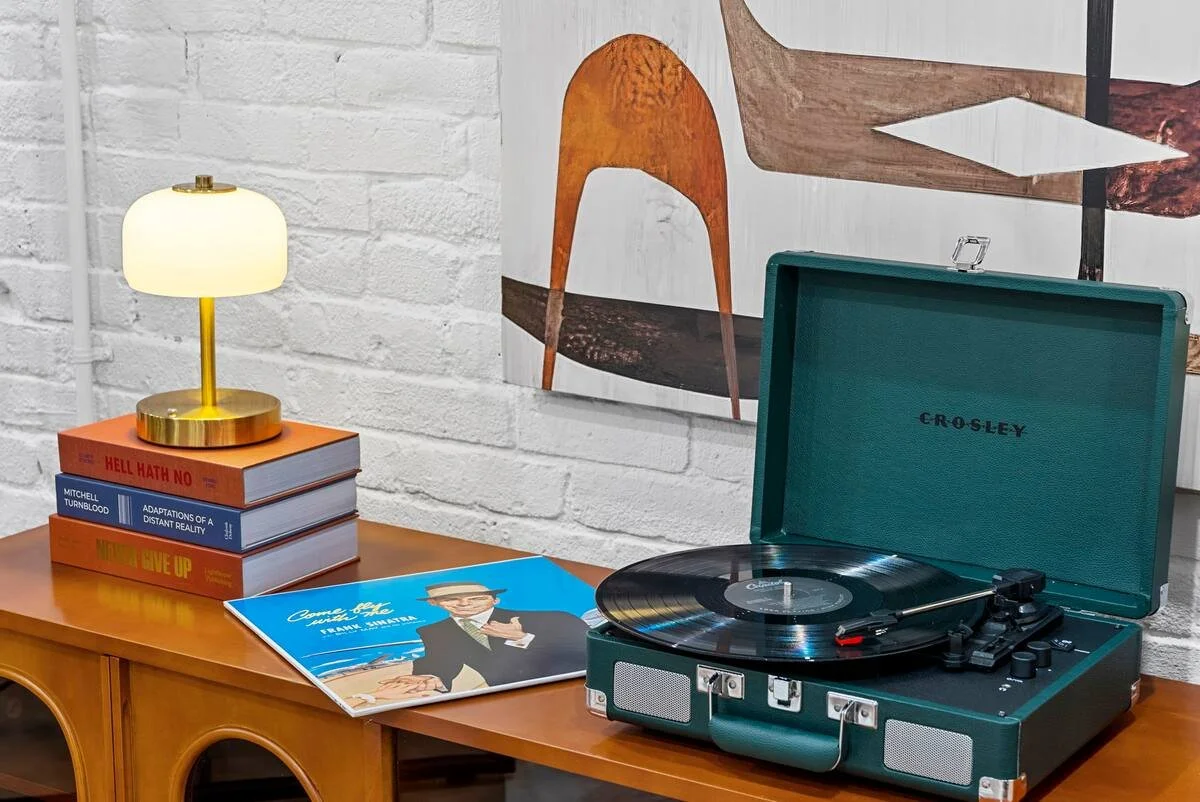 A vintage Crosley record player on a wooden sideboard, with vinyl records, a lamp, and a framed artwork on a white brick wall behind.