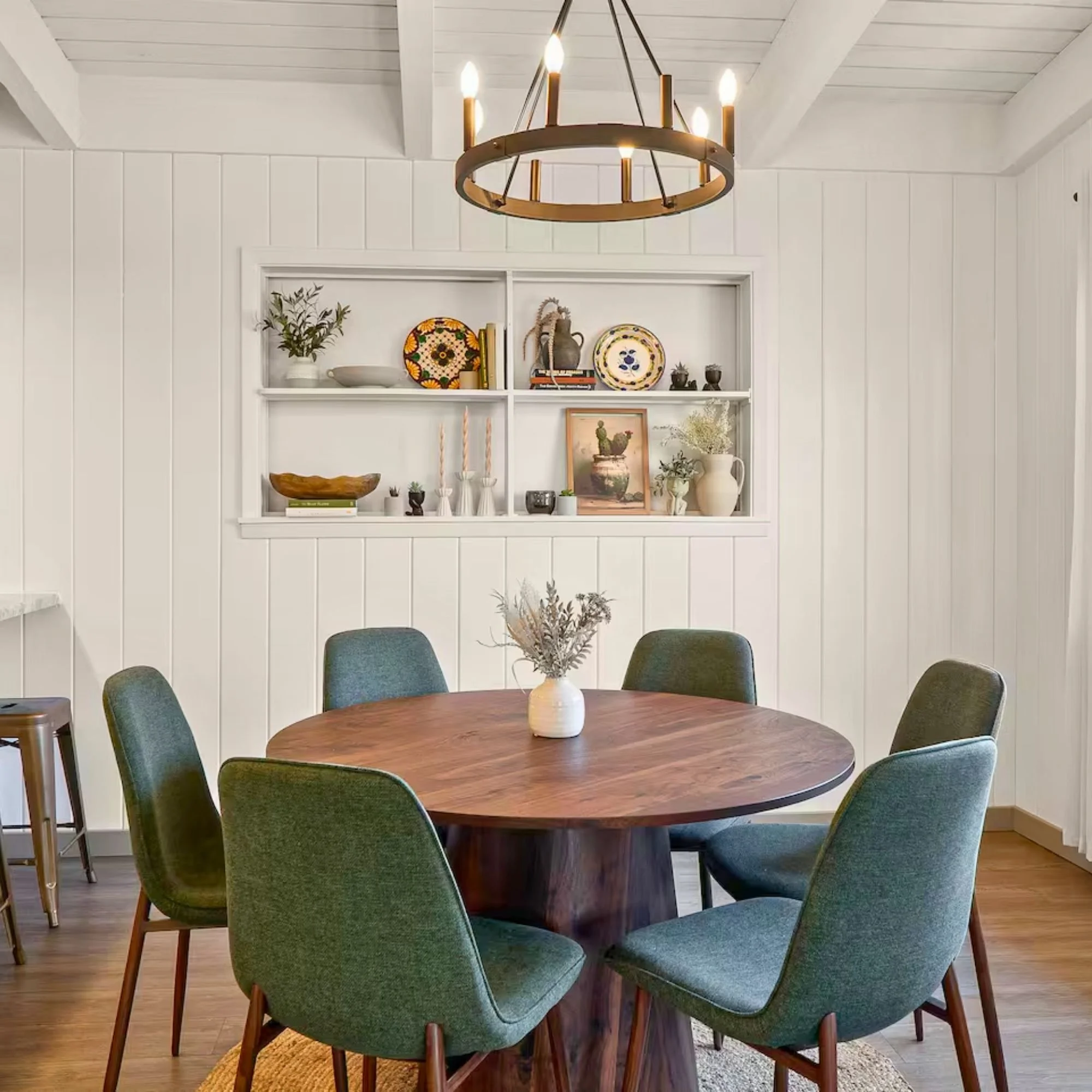 A round wooden dining table with a white vase holding dried flowers in the center. Six green upholstered chairs surround the table. In the background, white paneled walls with a built-in shelf displaying decorative plates, vases, books, and framed ar