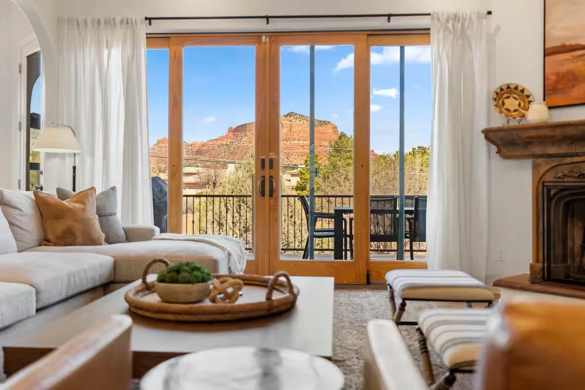 Living room with white sofa and pillows, coffee table with decorative bowl, glass sliding doors opening to a balcony with outdoor furniture, mountain view with red rock formation in the background, and white curtains framing the scene.