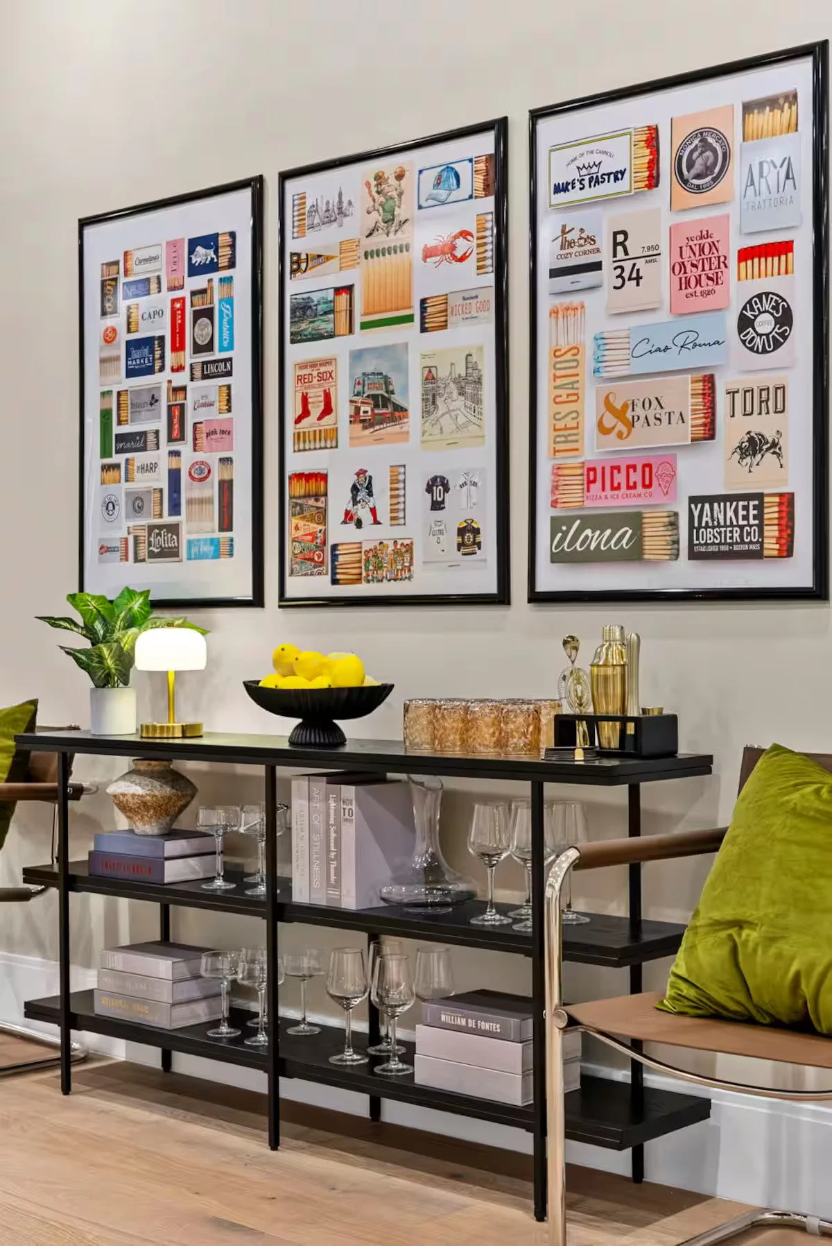 Decorative wall art displaying various matchbooks, framed and hanging on a wall. Below is a black shelf with glassware, books, a small lamp, and decorative items, against a light-colored wall.