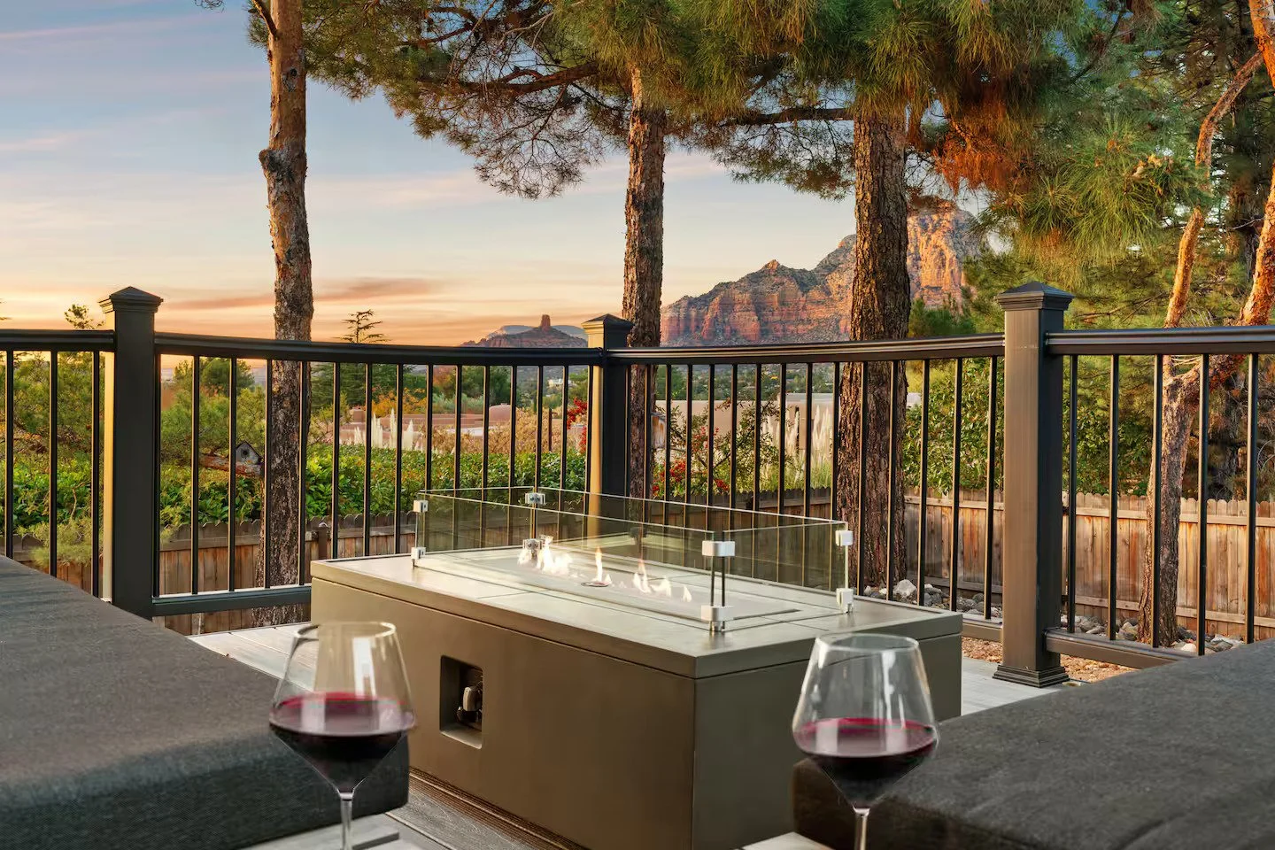 Outdoor deck with glass-enclosed fire pit, two glasses of red wine, and a scenic mountain view at sunset, surrounded by tall pine trees and a wooden fence.