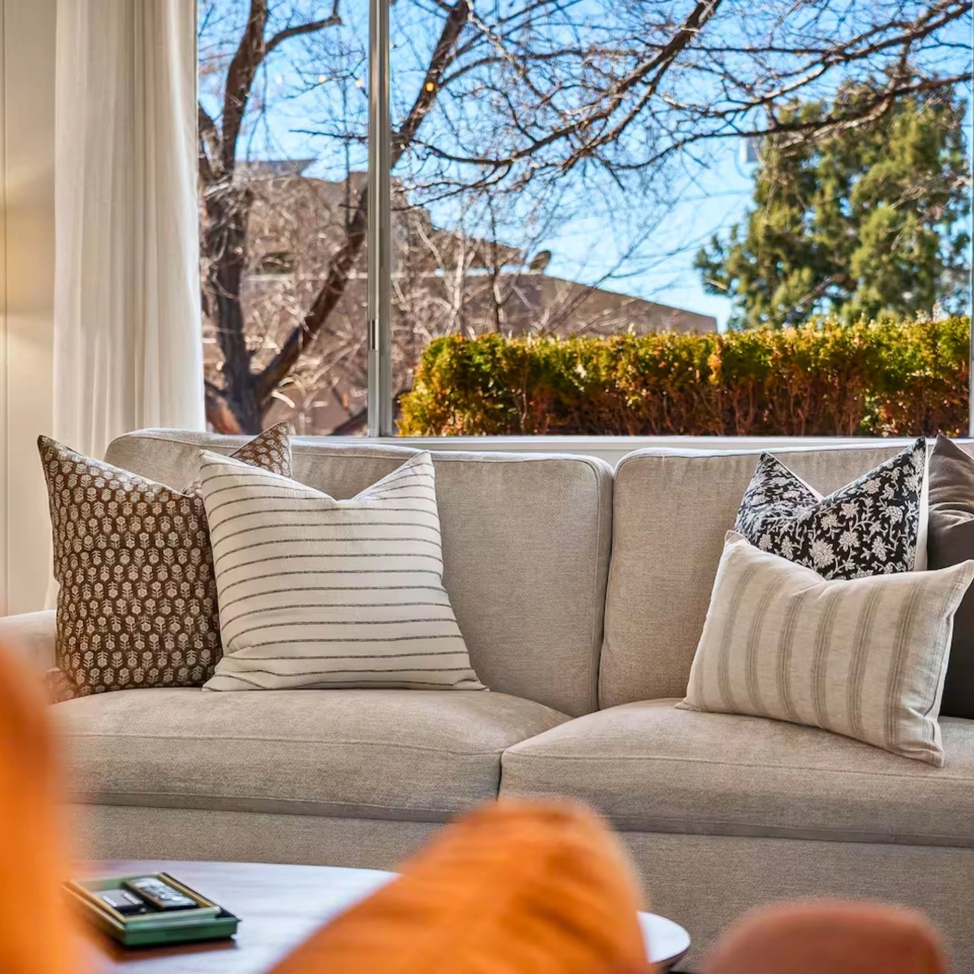 A living room with a beige sofa, decorative pillows, large window showing trees and a blue sky outside, and a small table with remote controls in the foreground.
