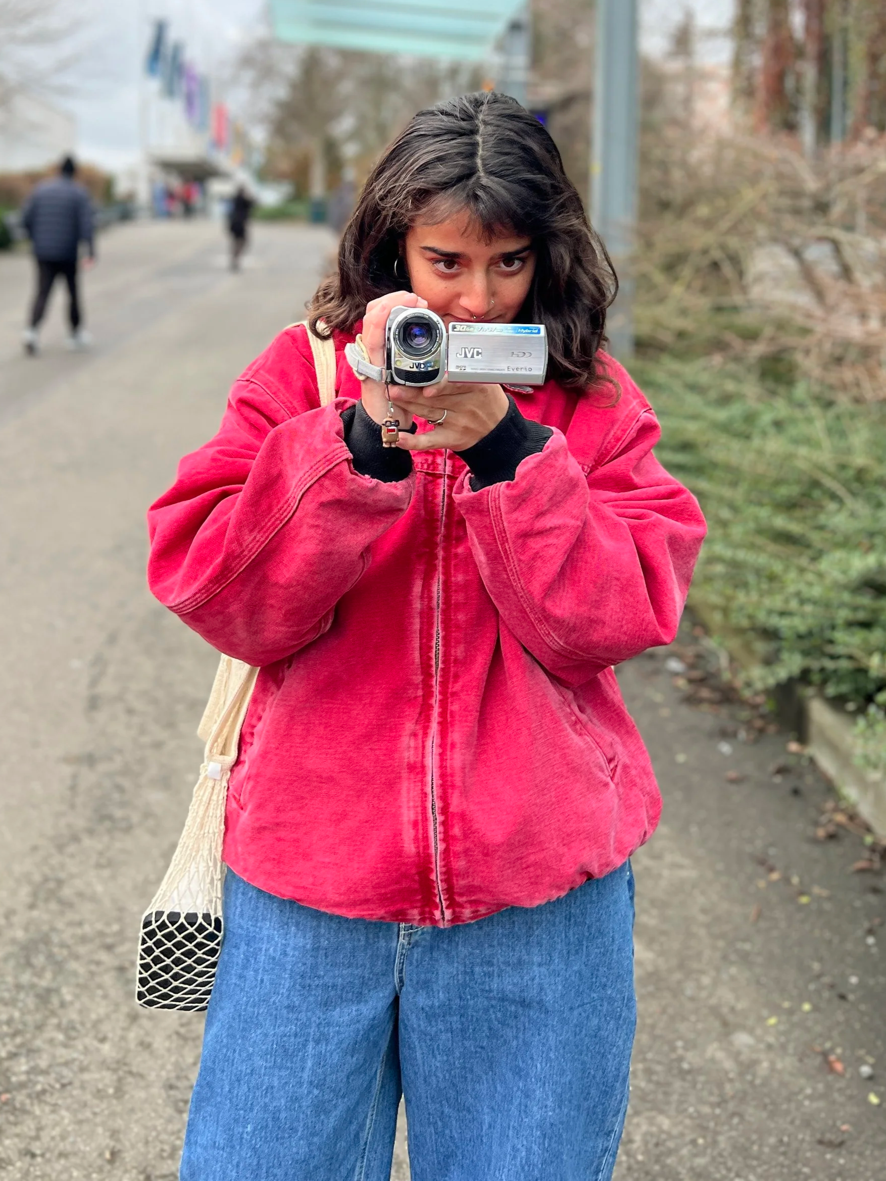 Young woman with dark hair and a septum piercing wearing a red jacket and blue jeans holding a camcorder pointed towards the camera, standing on a sidewalk outside.