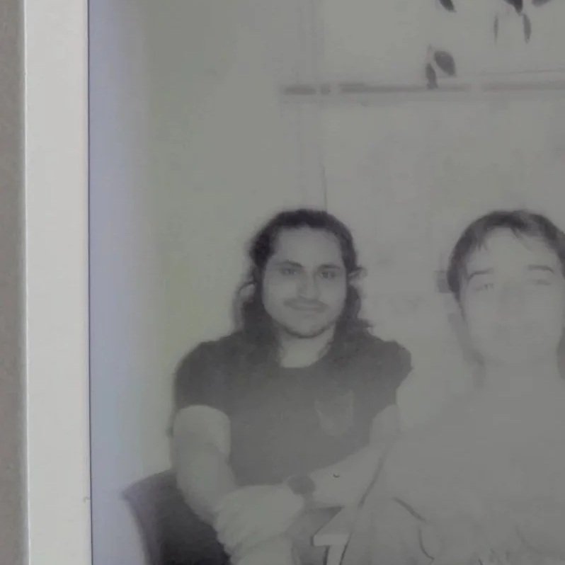 Black and white photo of a man with long hair, mustache, and beard, sitting with crossed arms, smiling.