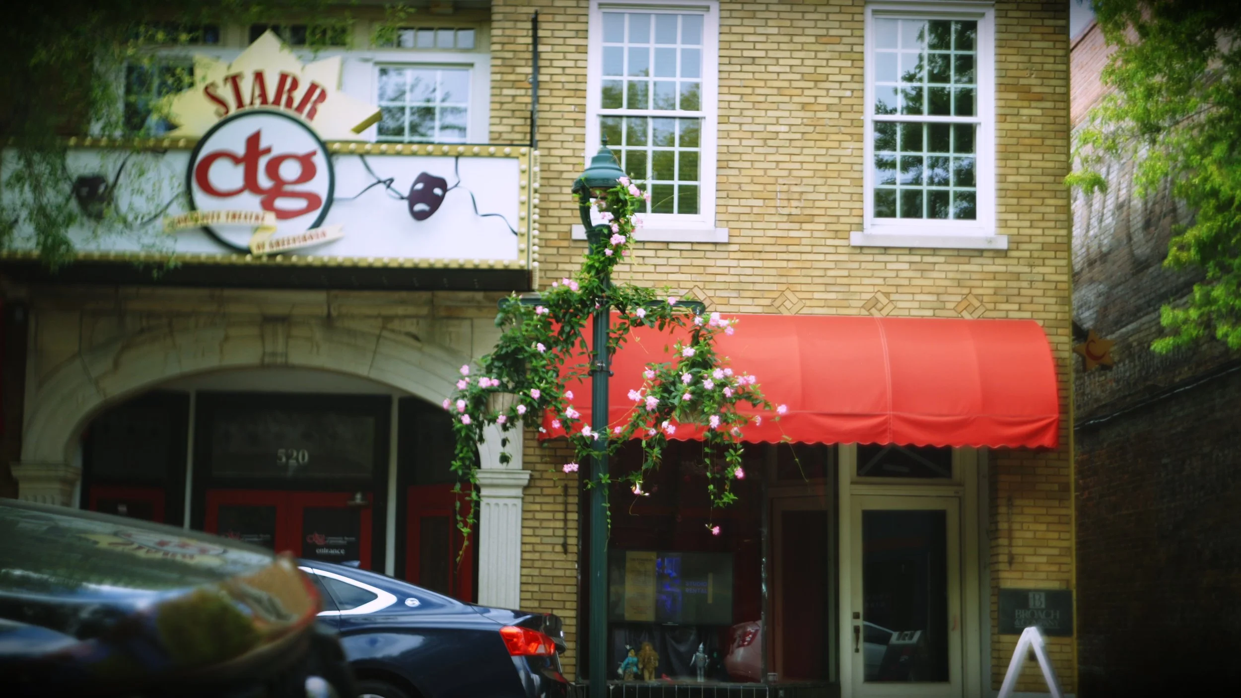 A street view featuring a brick building with a red awning, a decorated street lamp with pink flowers, parked cars, a sign for a theater, and two windows on the building facade.