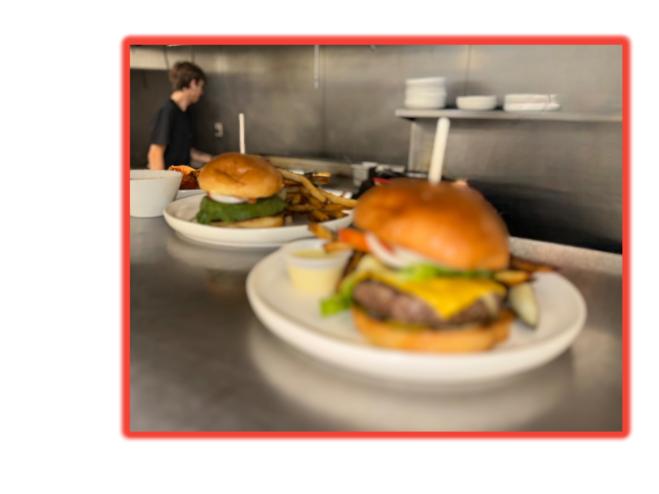 Two plates of burgers with fries and dipping sauce on a kitchen counter, with a woman working in the background.