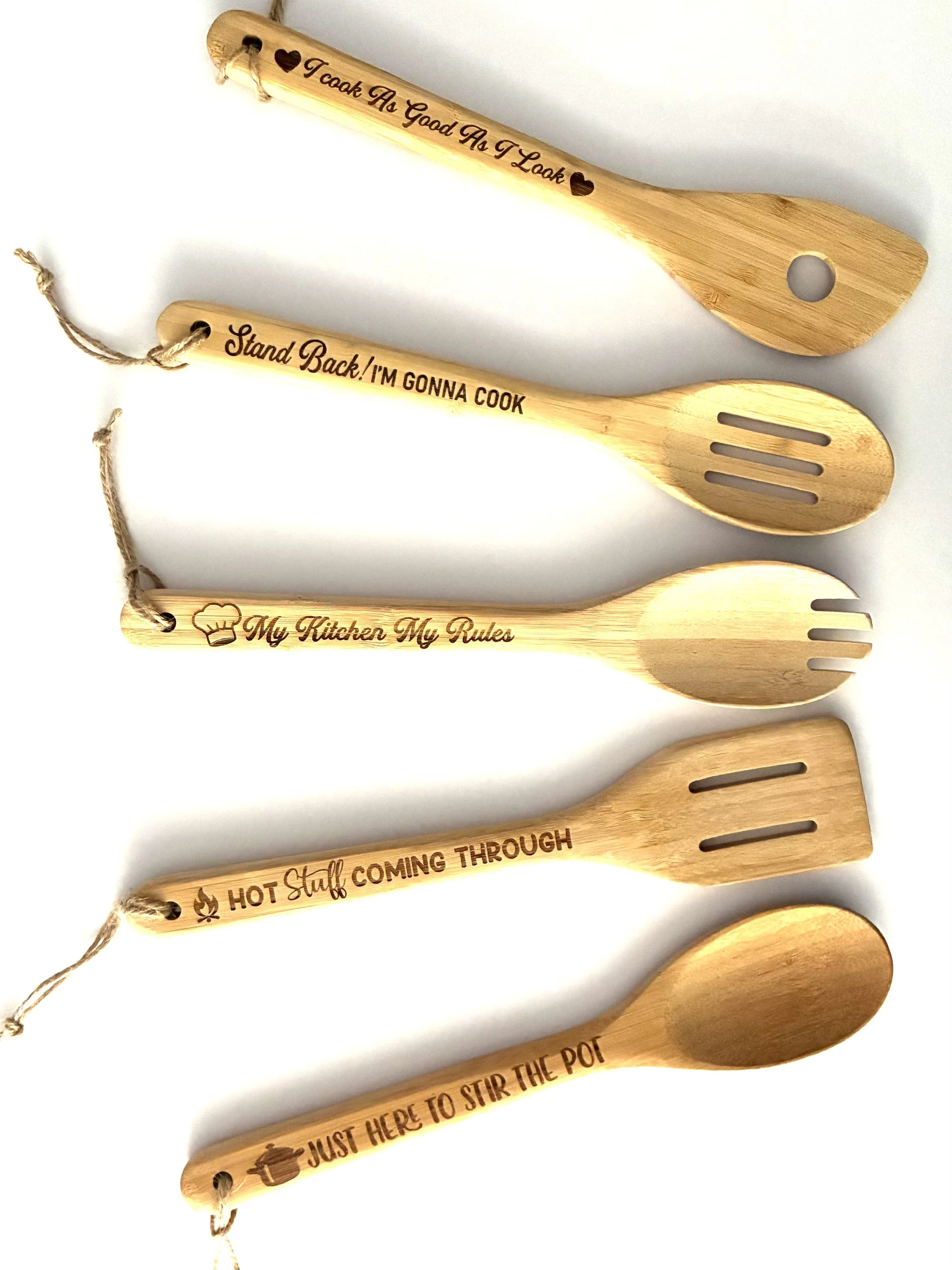 Five wooden kitchen utensils with engraved phrases and symbols, including a spatula, slotted spoon, and ladle, arranged in a row on a white background.