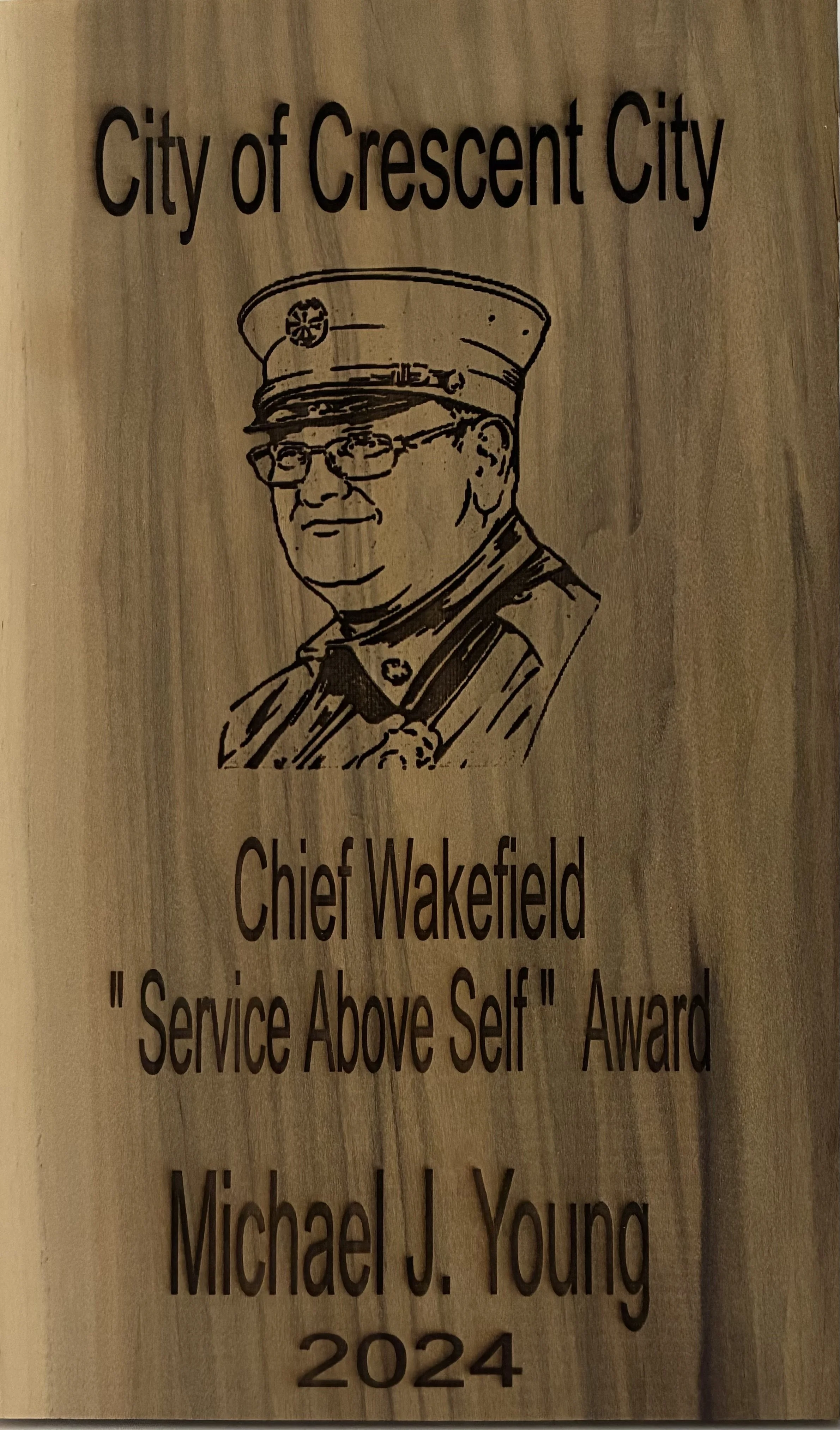 Wooden plaque with a drawing of a firefighter in uniform, an award from the City of Crescent City, awarded to Chief Wakefield for 'Service Above Self' in 2024, presented to Michael J. Young.