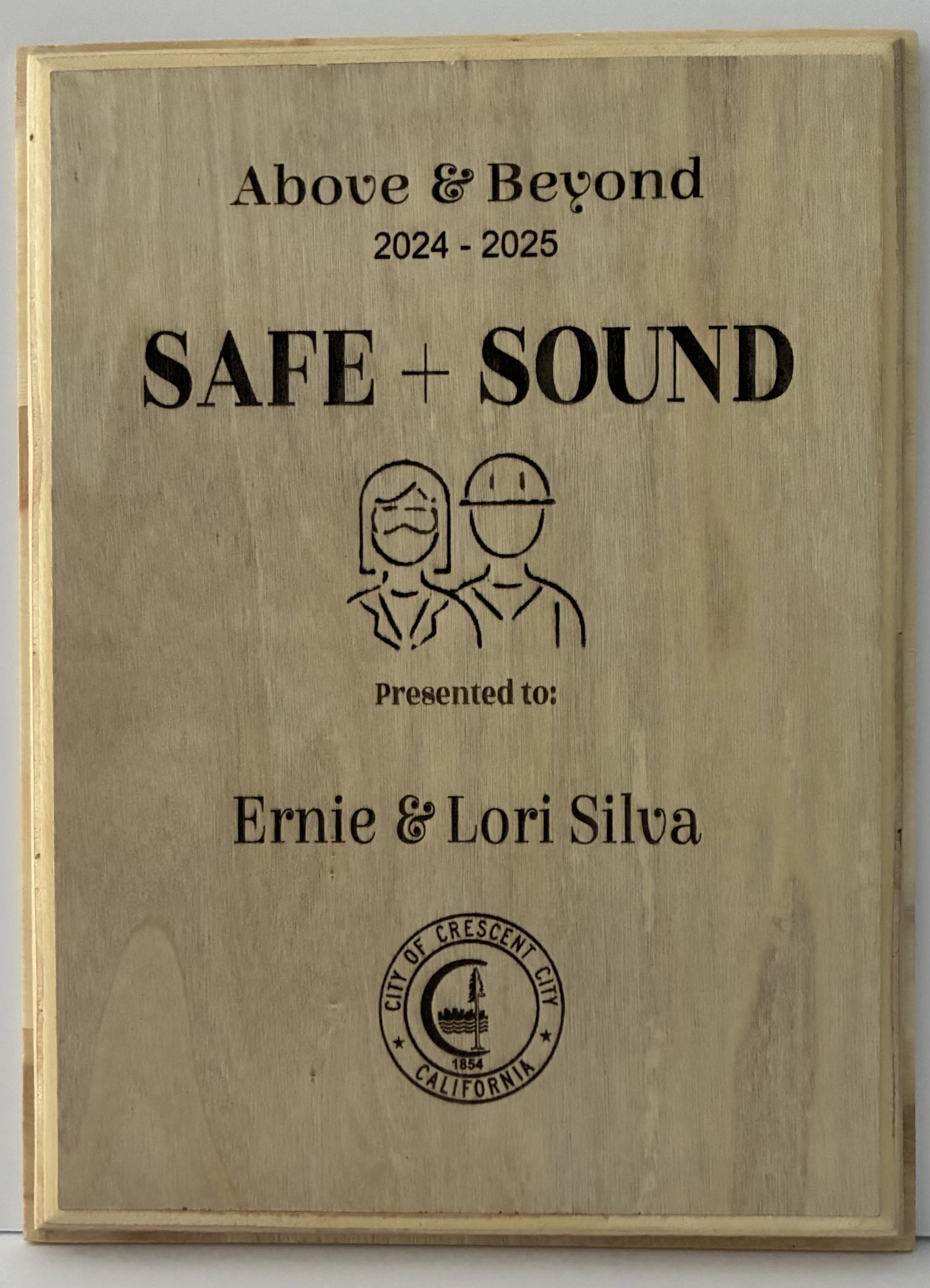 Wooden plaque with text and illustration. The text reads "Above & Beyond 2024 - 2025 SAFE + SOUND presented to: Ernie & Lori Silva". There is a simple line drawing of a smiling woman with glasses and a smiling man with a cap. At the bottom, the seal