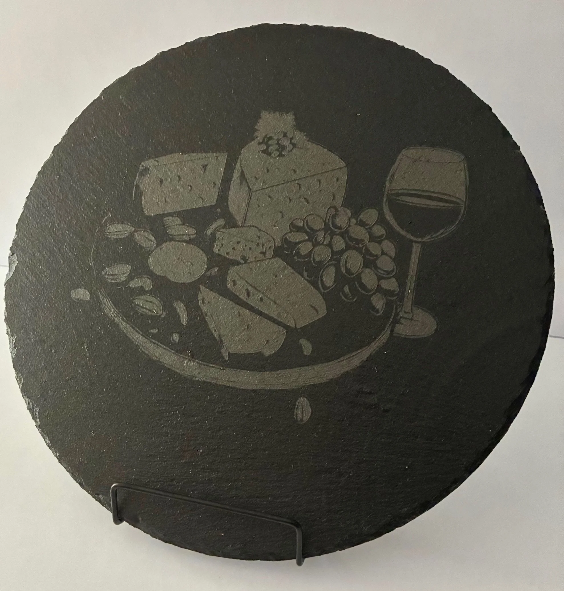 A round black ceramic plate with various cheeses, grapes, crackers, and a glass of red wine.