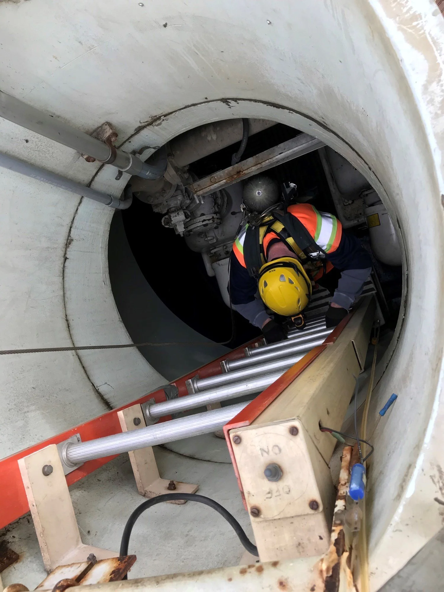 A worker wearing a yellow safety helmet, orange and safety vest, climbing a metal ladder inside a confined industrial space, surrounded by pipes and machinery.