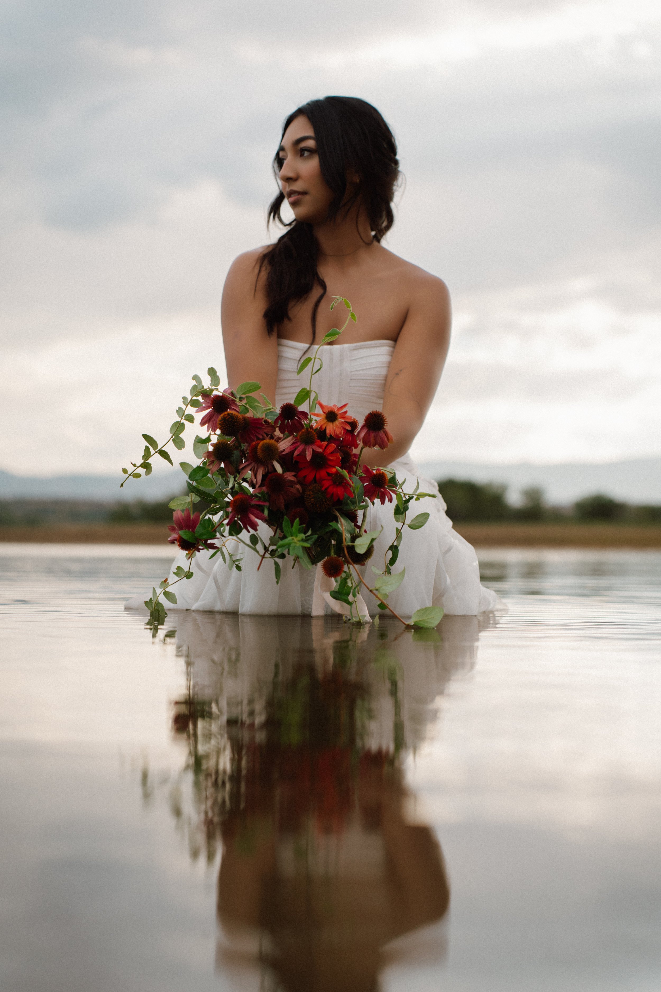 A person in a wedding dress kneeling in shallow water holding a bouquet of flowers