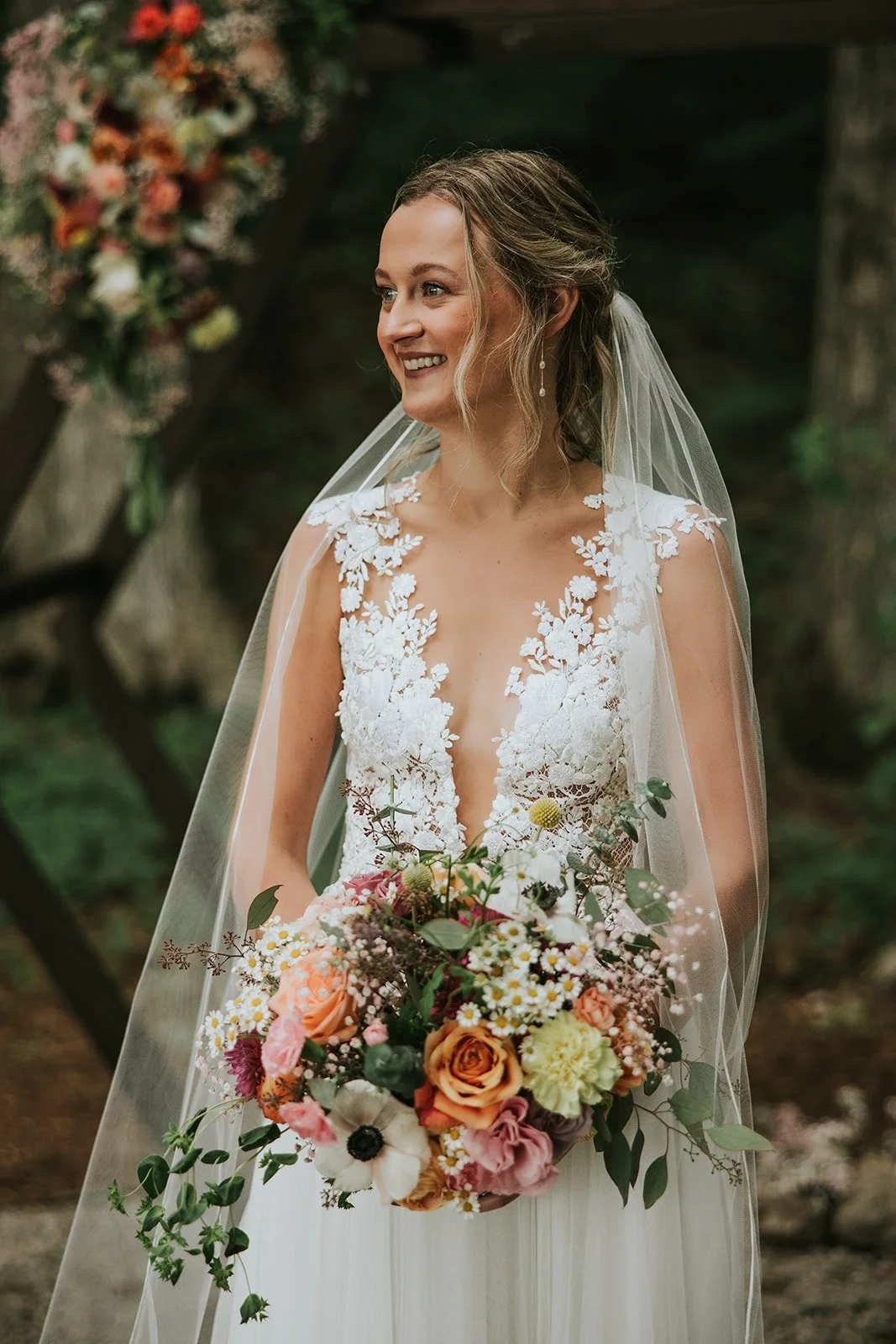Bride holding an Ash + Ether bouquet at a Colorado outdoor wedding, with warm-toned florals and a decorated arch behind her.
