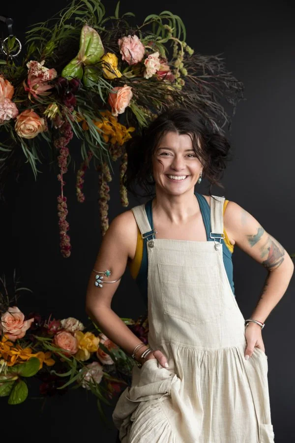 Ash smiling with her hands in her pockets as she stands next to floral arrangements