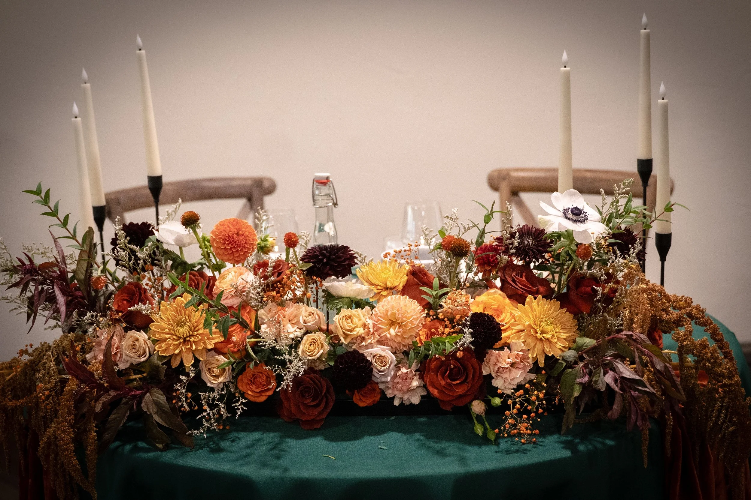 A table decorated with a large colorful floral arrangement and four tall white candles in black holders, with two wooden chairs in the background.