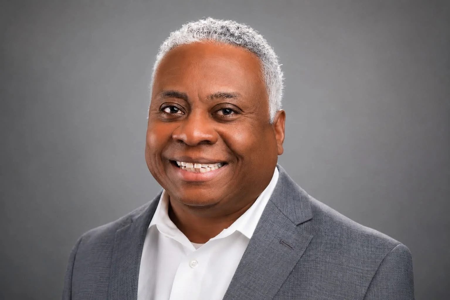 A portrait of a smiling African American man with short curly gray hair, dressed in a navy blue plaid suit, a light blue dress shirt, and a green striped bowtie, against a plain light background.