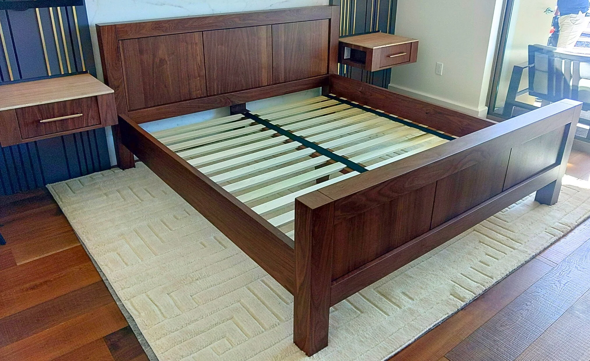  Wooden bed frame with headboard and side rails in a room with a beige and brown rug and two small wooden side tables attached to the wall.