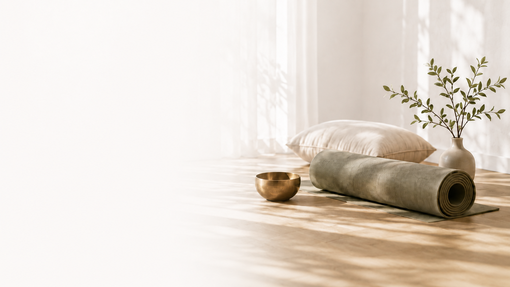 Yoga mat, pillow, brass singing bowl, and vase with green leaves on a wooden floor near a window