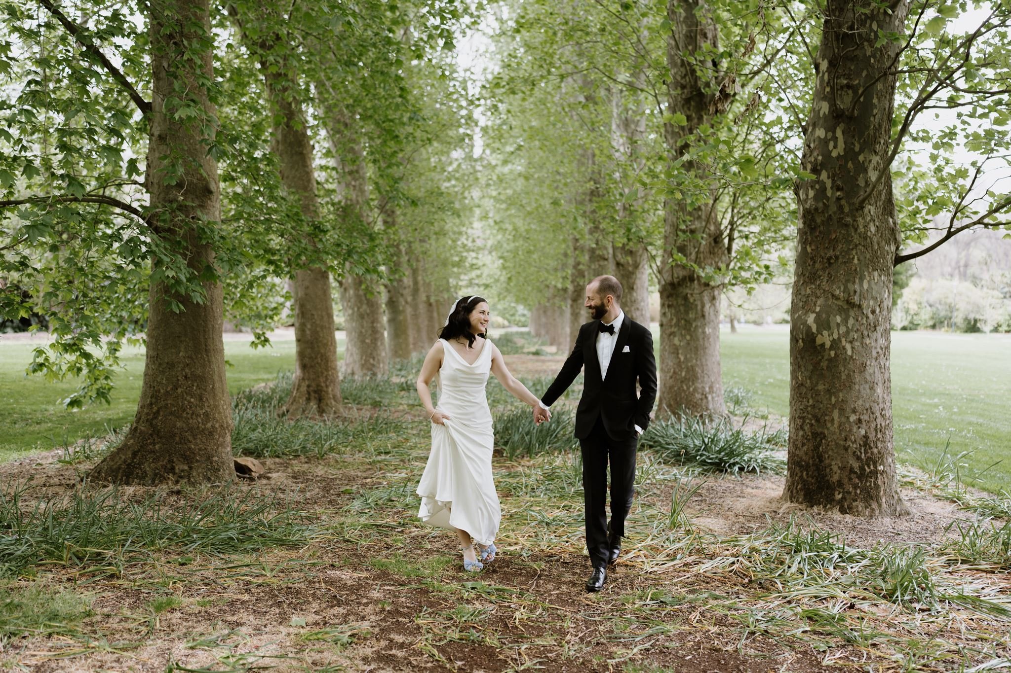 A bride and groom walking hand in hand through a park with tall trees and lush greenery, smiling at each other.